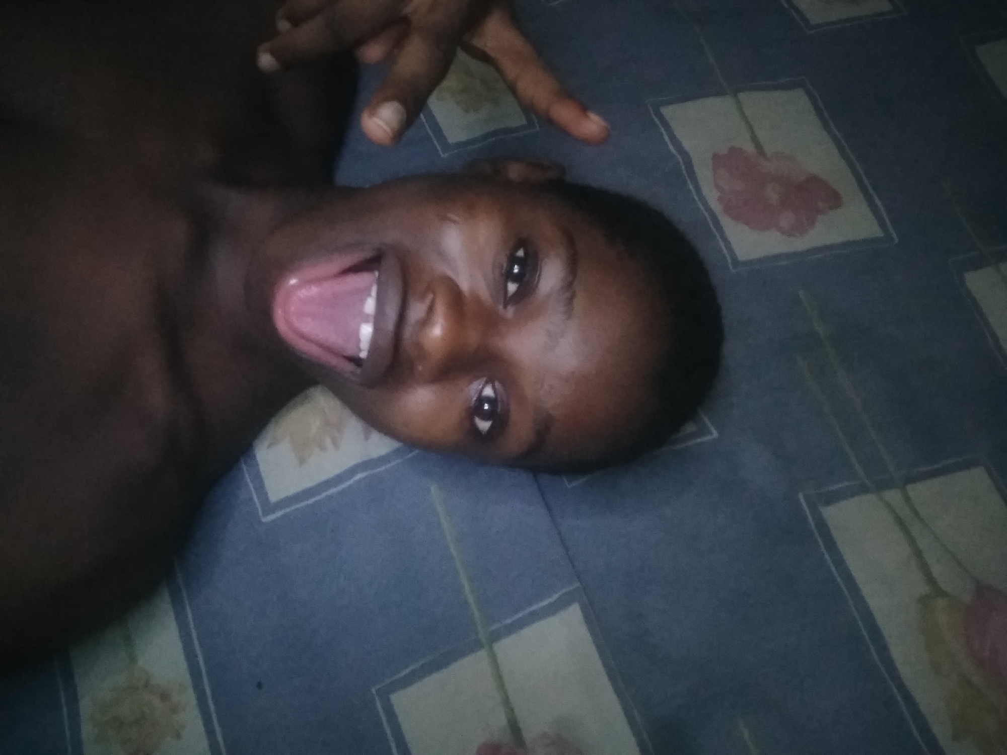 A dark-skinned person is lying on a bed with a patterned blue and white bedsheet. They are facing upwards, with their mouth open and tongue visible. One hand is visible above their head, displaying two fingers. The location is presumed to be Shangisha, Nigeria, but this cannot be confirmed from the image.