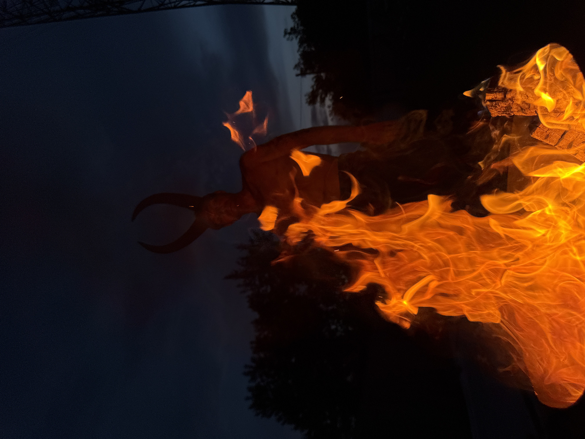 A person with horns is silhouetted by a fire at dusk, with a dark sky and trees in the background. The individual appears to be shirtless and is positioned behind a large, vibrant fire. The fire is the primary light source, casting an orange glow on the person and the surrounding area. The environment suggests an outdoor setting, possibly a backyard or park, given the presence of trees and the dark, twilight sky. The location of Chaikovsky, Russia is not discernible from the image.