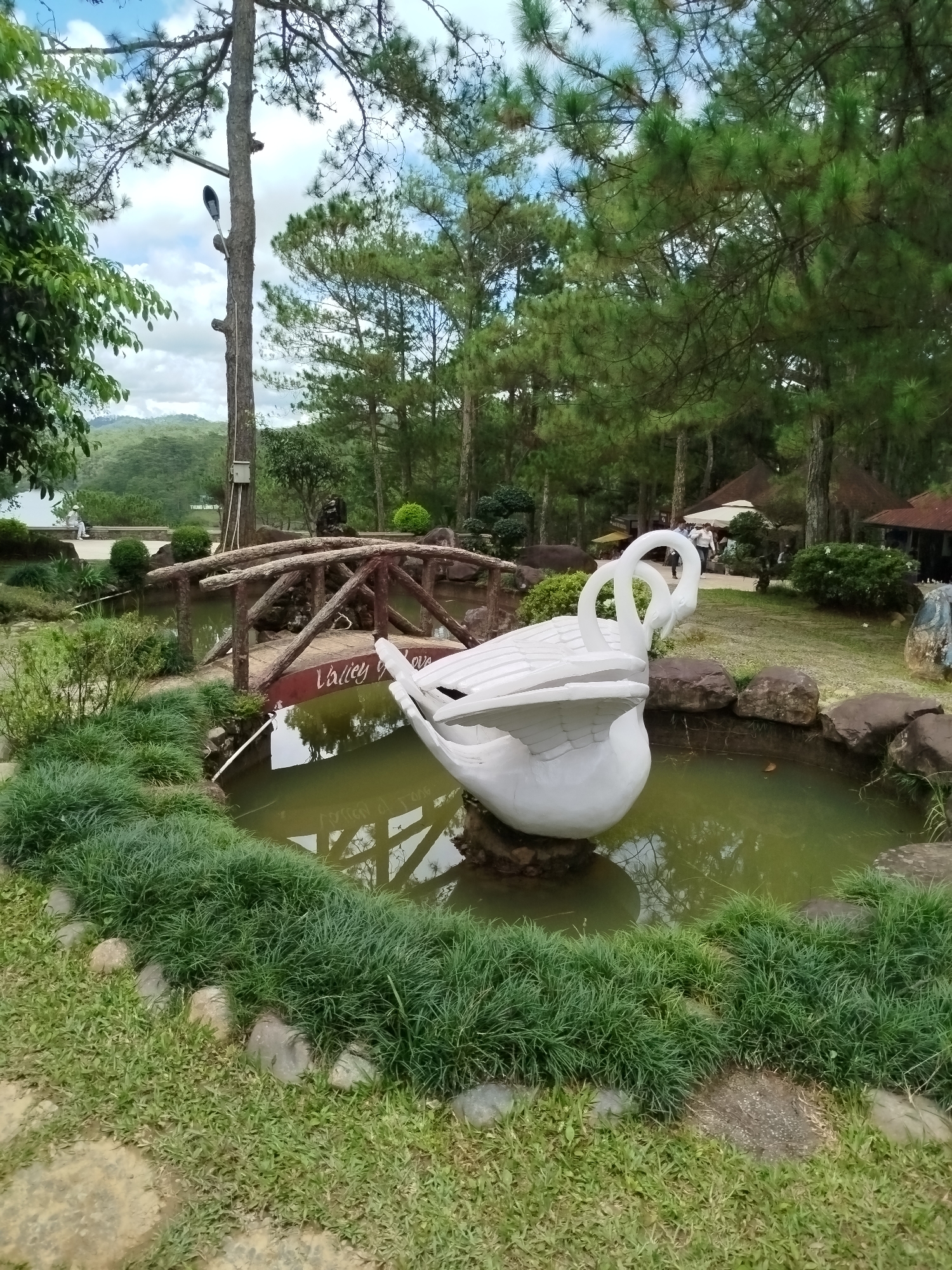 The image depicts a serene park scene at Lâm Viên - Đà Lạt, Vietnam. In the foreground, a large, white swan sculpture is positioned in a pond, with its neck arched gracefully. The pond is surrounded by lush green grass and rocks. A rustic wooden bridge spans a section of the pond, and on its surface, the words "Valley of Love" are visible, indicating the park's name. In the background, tall pine trees dominate the landscape, with a hint of a lake and rolling hills in the distance. The sky is partly cloudy. There are a few people in the background, walking near some of the park's structures, which appear to be covered by thatched roofs. A lamppost is attached to a tall tree on the left side of the frame. The overall impression is one of natural beauty and tranquility.