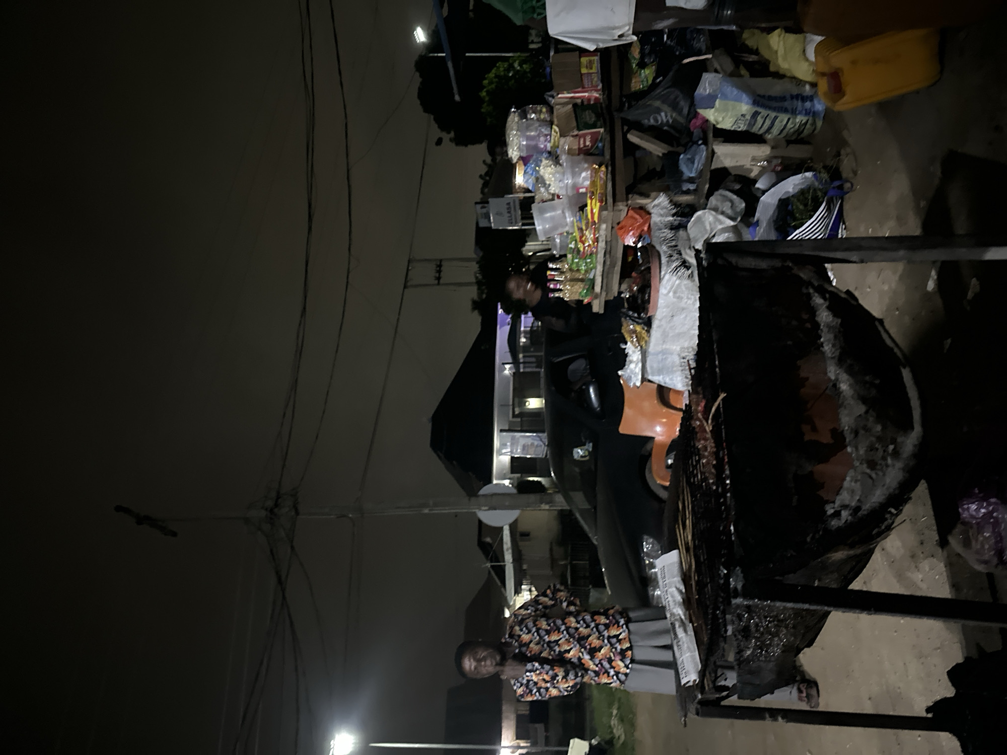 At night, on a dusty, outdoor street in Abuja, Nigeria, a street vendor is attending to a grill filled with cooked food. The vendor, a man wearing a colorful patterned shirt and gray pants, stands to the left of the grill. To the right of the grill, a woman is standing behind a table laden with various goods, including snacks, drinks, and other packaged items. The table is covered with a white cloth, and various bags of produce and other items are stacked behind it. A dark-colored car is parked behind the table. The scene is illuminated by a street lamp and possibly other ambient lights. In the background, buildings and utility poles are visible against the dark night sky.