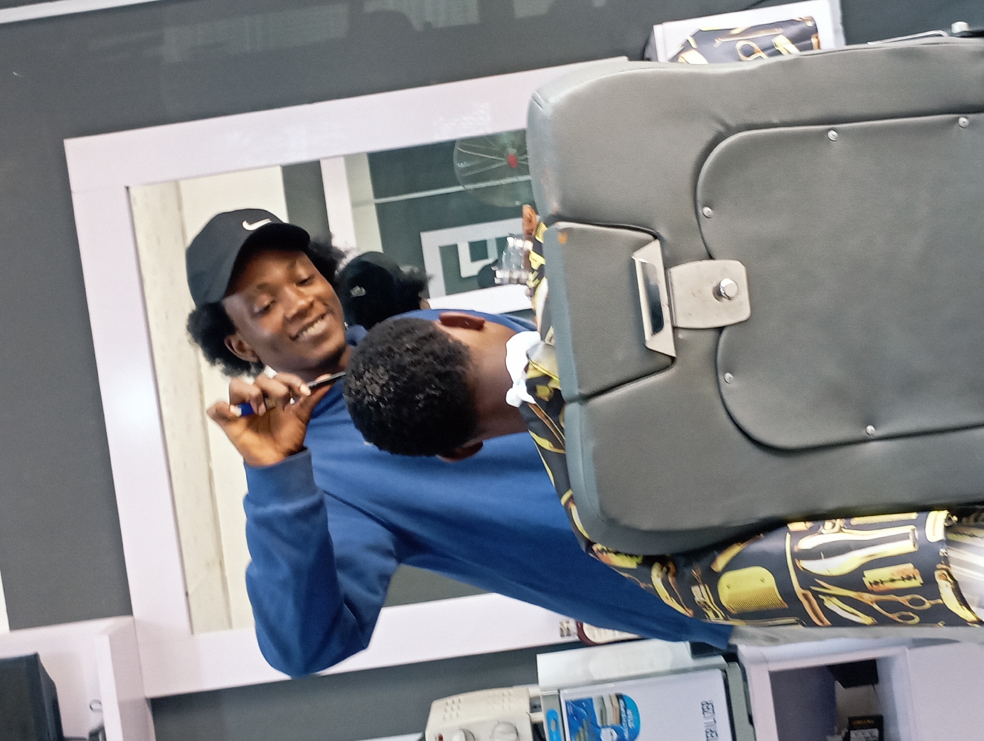 The image captures a dynamic scene within a barber shop, likely in Jos, Nigeria, featuring a barber attending to a client. The photograph is taken at an angle, with a large mirror dominating the background, reflecting parts of the shop and the individuals.
The primary subject is a young male barber with dark, curly hair, wearing a black Nike baseball cap and a blue long-sleeved shirt. He is smiling broadly, holding a barber's tool (possibly a razor or comb) in his hand, and appears to be actively working on the client. His reflection, showing his smiling face, is clearly visible in the mirror.
The client, also a young male, is seated in a sturdy, grey upholstered barber chair. His head and upper body are visible, with short, dark, curly hair. White shaving cream or foam is applied to the side of his head near his ear, indicating he is receiving a shave or a precise haircut detail. A distinctive barber's cape or cloth, patterned with gold scissors, razors, and combs on a dark background, is draped over him and the chair.
The activity central to the image is professional barbering, with the barber meticulously styling or shaving the client. The barber's cheerful expression suggests a positive and engaging atmosphere.
Notable details include the professional setup of the barber shop, complete with a large, white-framed mirror. In the reflections, a ceiling fan with red blades can be faintly seen. On a shelf below the mirror, various items are visible, including what appears to be a white electronic device and a blue-labeled product where the word "SPLITTER" is partially legible. The overall setting points to a well-equipped and active grooming establishment.