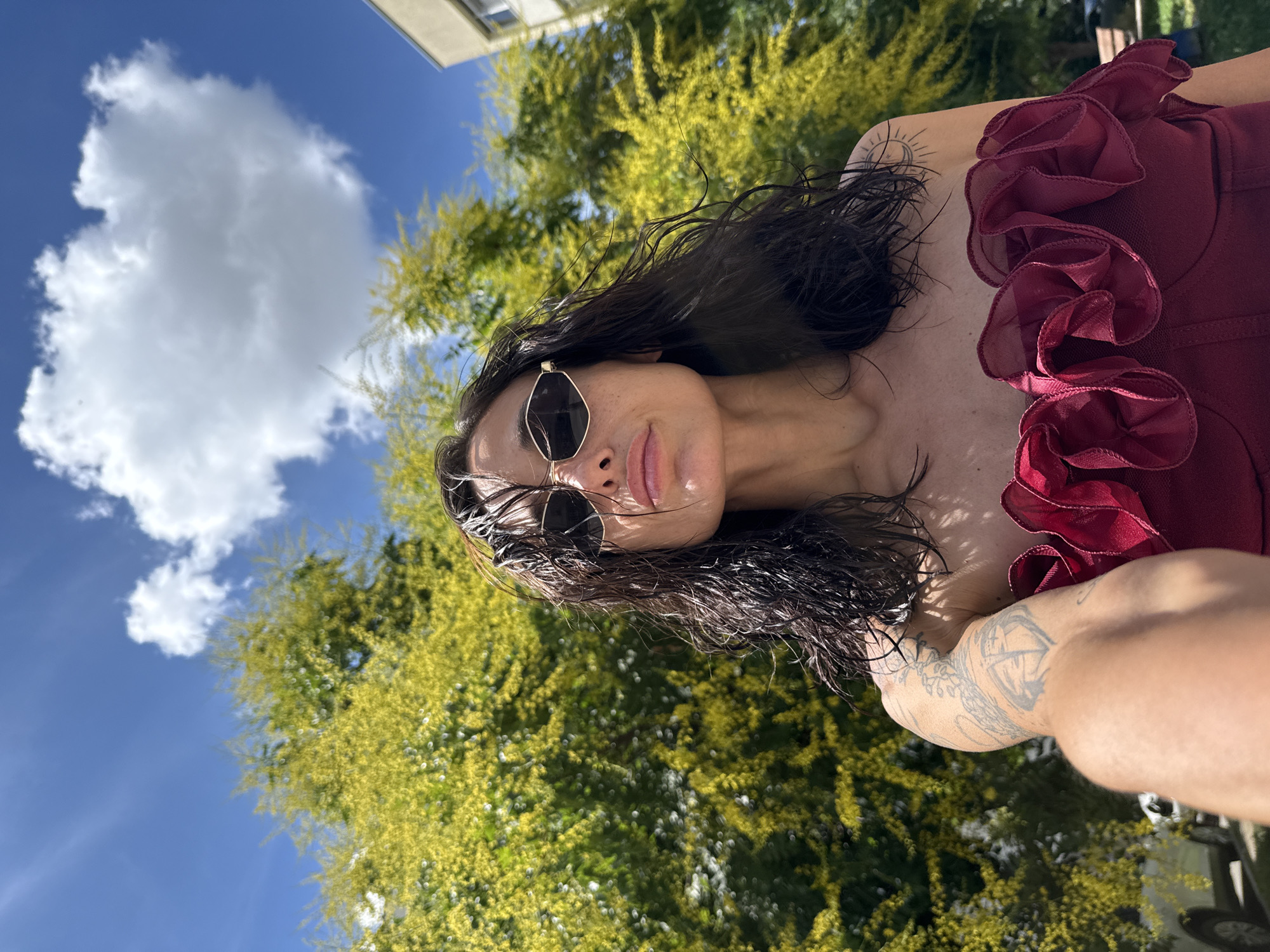 The image is a selfie taken outdoors in Pleven, Bulgaria, under a bright blue sky with a large white cloud. The subject is a woman with dark, wet hair, wearing sunglasses with gold frames. She is wearing a dark red strapless top with ruffled detailing around the neckline. Her left arm is visible, showing tattoos. Behind her is a large tree with vibrant green and yellow foliage, and a portion of a light-colored building is visible on the right side of the frame. A white car is partially visible in the lower left corner. The overall impression is that of a warm, sunny day, possibly during summer.