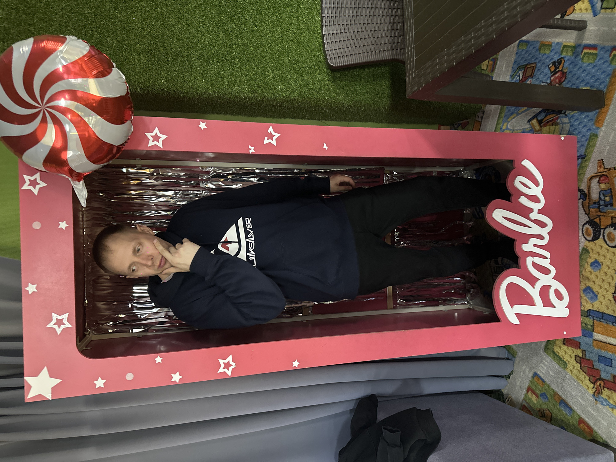 A man is posing inside a large, pink "Barbie" themed frame. The frame is decorated with white stars and the word "Barbie" in white cursive. The interior of the frame is lined with silver, reflective material. The man is wearing a dark blue hoodie with a white logo and black pants. He is holding his hand to his face in a contemplative pose. Next to the frame is a red and white swirl balloon. The scene is located indoors; a brown wicker table and chairs sit on a green, artificial lawn in the background, and a colorful rug is visible near the frame, alongside a gray couch. According to the metadata, the location is Glazov, Russia.