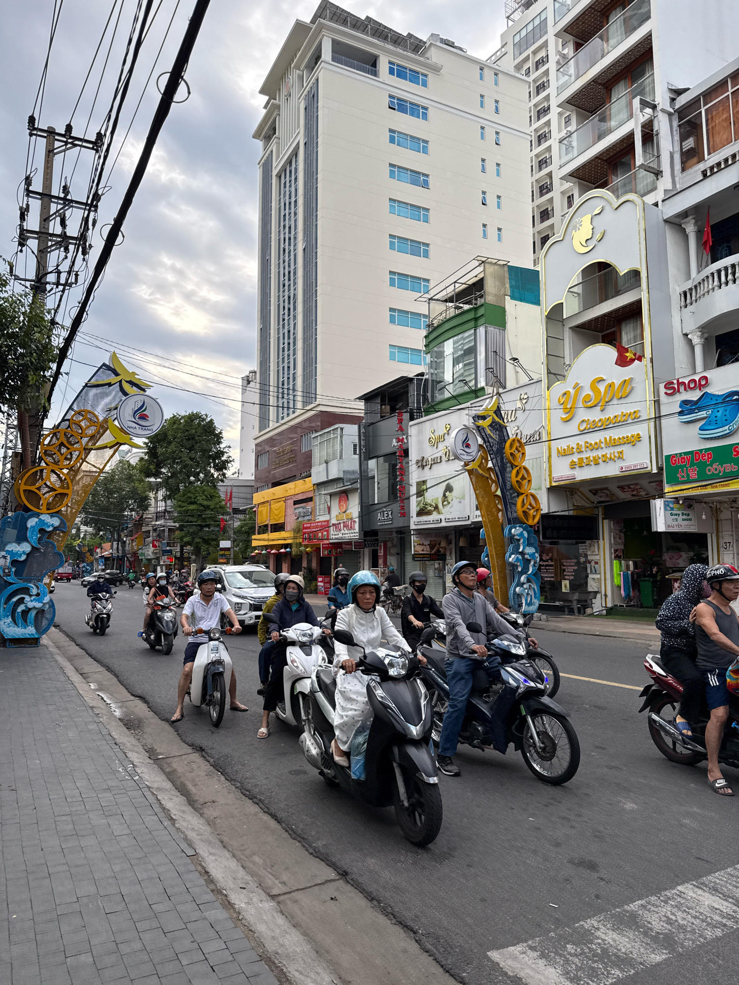 The image captures a street scene in Nha Trang, Vietnam, likely during daytime, under a cloudy sky. The foreground features a busy road with numerous motorbikes and riders, indicating active city traffic. The people on the motorbikes are dressed in casual clothing, with some wearing helmets for protection. The motorbikes themselves are varied in make and model, commonly seen in Southeast Asian cities.
On the right side of the road, a row of commercial buildings lines the street. These buildings appear to be multi-story structures with storefronts at street level. One prominent sign displays "Y Spa Cleopatra," advertising beauty and massage services, with Vietnamese text also visible. Another sign depicts a pair of blue flip-flops with the word "Shop" and "Giày Dép," suggesting a footwear store. The architecture of these buildings is modern, with glass windows and balconies.
The left side of the street shows a pedestrian walkway paved with interlocking bricks. Adjacent to this is a large decorative archway with a circular emblem that reads "NHA TRANG" and features a symbol of a bird or phoenix. The archway is illuminated with yellow lights and decorated with blue wave-like motifs and circular elements. Beyond the archway and walkway, trees and more buildings are visible, contributing to the urban landscape.
In the background, taller buildings, including a modern white high-rise with many windows, dominate the skyline. Overhead, a complex network of electrical wires and utility poles crisscrosses the sky, a common sight in many Asian cities. The overall impression is one of a bustling urban environment in a coastal city, with a blend of commercial activity, everyday life, and prominent local signage. The presence of Vietnamese flags on some buildings further confirms the location.