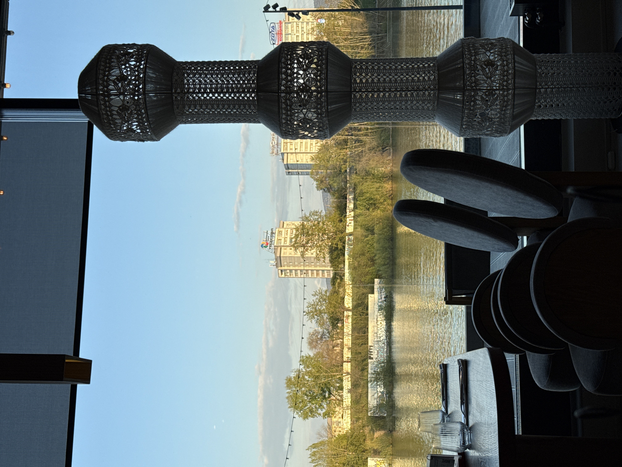 The interior of a cafe with a view of the Yenisei River in Krasnoyarsk, Russia. A woven decorative lighting fixture is suspended from the ceiling. A window with a dark shade pulled down partially reveals an exterior view. The Yenisei River reflects the sky and the city buildings along the bank. A row of buildings is visible in the background and along the river bank. Seating for patrons is visible in the foreground, including several chair backs and table settings. The scene appears to be set during the day with natural light illuminating the interior.
