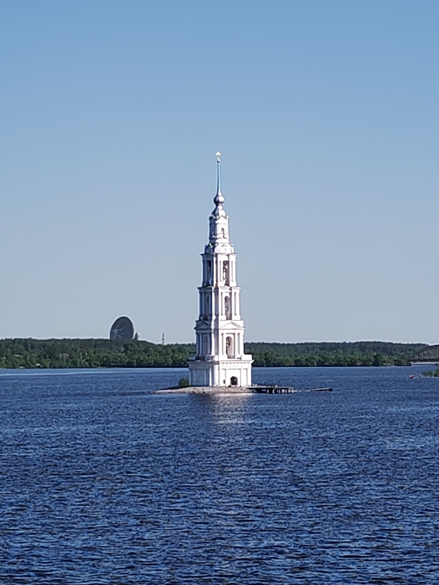 The image shows the flooded bell tower of St. Nicholas Cathedral in Kalyazin, Russia. The white, tiered bell tower stands prominently in the middle of a body of water, presumably the Volga River. A small island supports the tower, with a partially submerged dock or structure extending from it. The water has a dark blue, textured surface due to visible ripples. In the background, a shoreline with a dense forest is visible. To the left of the bell tower, a large satellite dish or radar structure can be seen behind the forest, partially obscured by the trees. The sky is clear and light blue.