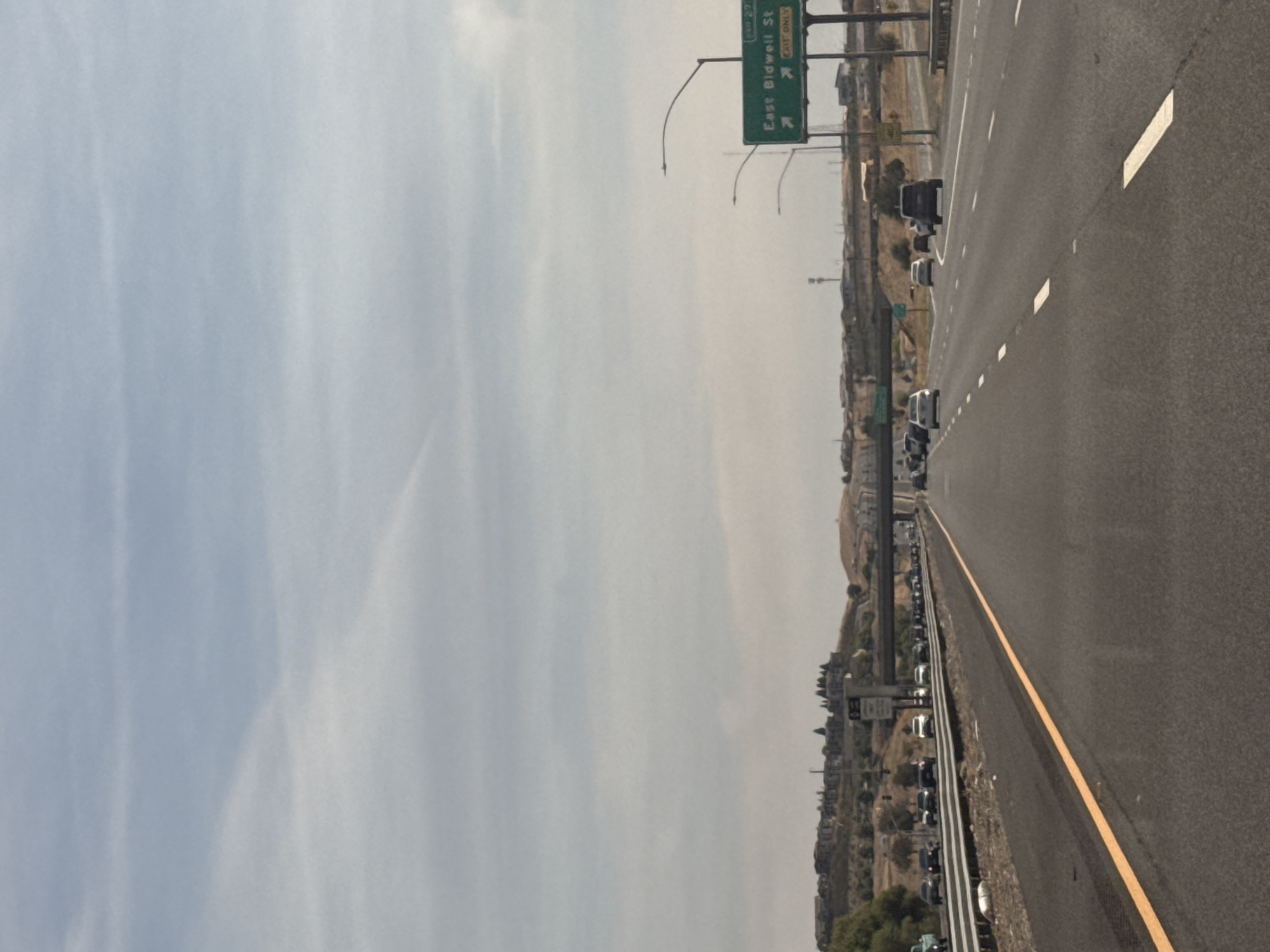 This is an image of a highway in Folsom, California, USA. The highway has multiple lanes of traffic, with cars traveling in both directions. There is a road sign indicating "East Bidwell St" and "Exit Only." The sky is overcast with clouds. In the background, there are hills with some buildings and trees. The image appears to be taken from a moving vehicle, as indicated by the slight blur and the perspective.