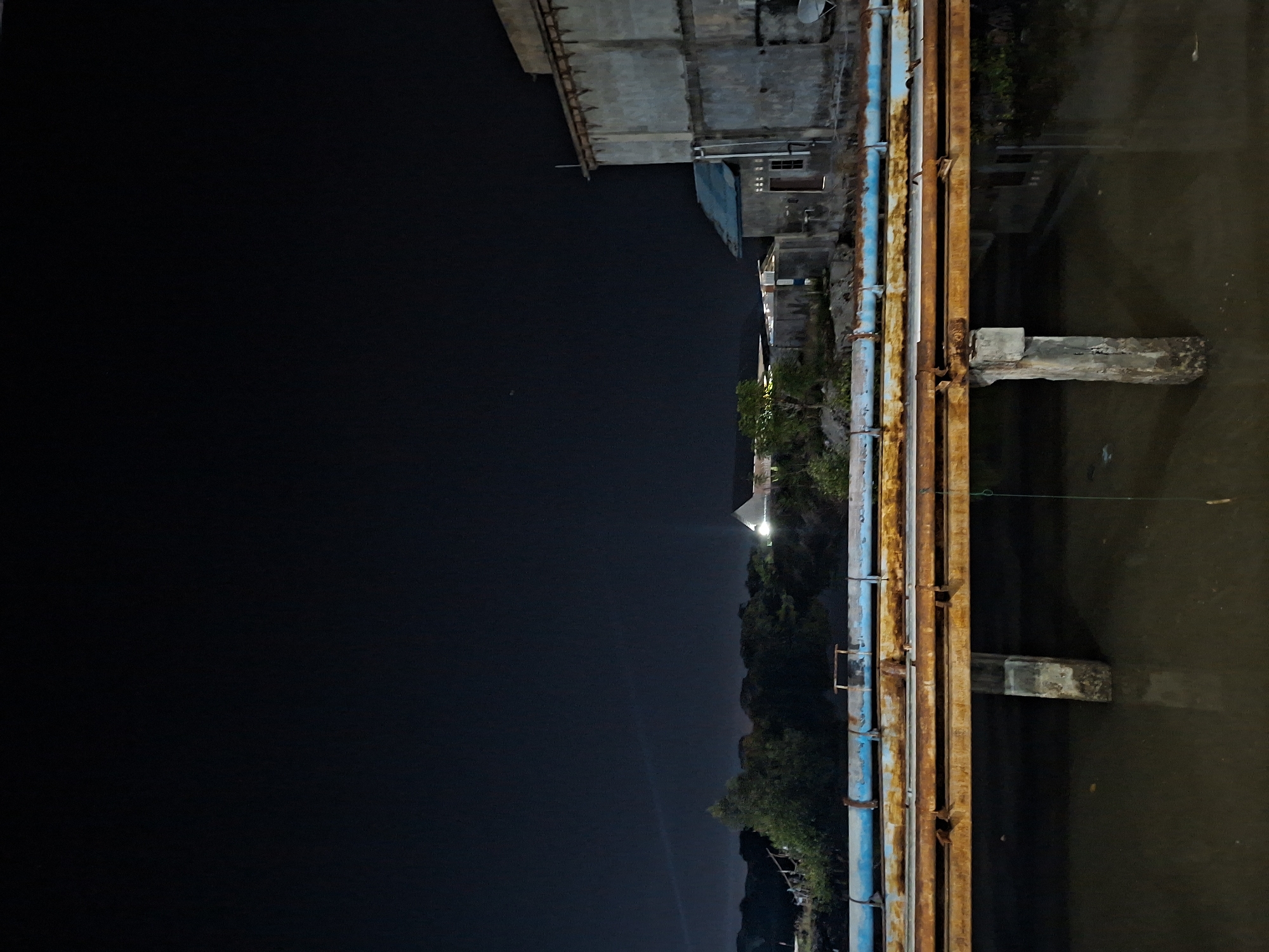 This nighttime photo shows a waterway in Lhokseumawe, Indonesia. Several parallel pipes, some blue and some rusted, run along the edge of the water, supported by concrete piers. Adjacent to the waterway are buildings, trees, and what appears to be a lit-up structure further in the distance. A substantial dark space occupies the left side of the frame, likely representing the night sky or a dark landmass.