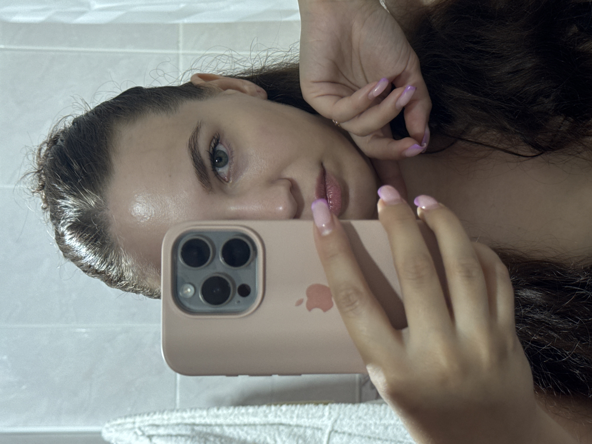 The image depicts a mirror selfie of an adult female. Only the right side of her face is visible, with her left eye, nose, mouth, and chin partially obscured by a light pink iPhone in a matching silicone case. She has dark, damp hair pulled back, and her visible right eye is light green or hazel. Her expression is neutral, and she looks directly into the camera.
Her left hand holds the iPhone, showing manicured nails with a pink base transitioning to purple tips. The iPhone case features a visible Apple logo and a prominent camera module with three large lenses and additional sensors. Her right hand rests against the right side of her jaw and neck, displaying similarly manicured nails and a thin, metallic ring on her ring finger.
The background consists of light-colored, rectangular tiled walls with grey grout, and a white textured towel is visible hanging on the left side of the frame. No identifiable landmarks or specific geographical indicators are present within the image to determine a precise location such as Chaikovsky, Russia.