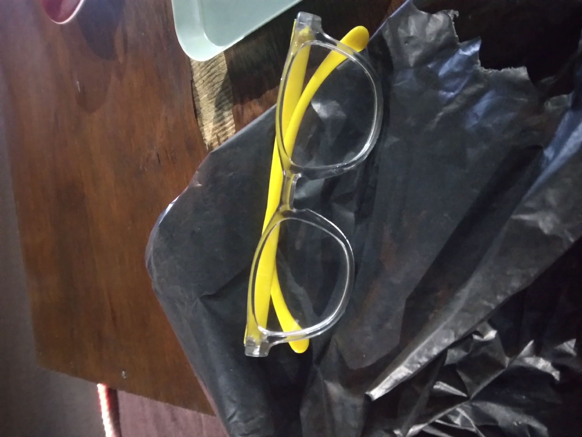 A pair of clear-rimmed glasses with bright yellow arms rests on a crumpled black plastic bag. The glasses are positioned diagonally, with the left arm pointing towards the top left of the frame and the right arm extending towards the bottom right. The background consists of a dark wooden surface, possibly a table, with subtle texture and grain. To the right of the glasses and bag, a light blue plastic object and a portion of a red saucer are visible on the wooden surface. The lighting appears dim, casting soft shadows. There are no people present in the image. The overall scene suggests an indoor setting, possibly a home or a cafe, in Pacet, Indonesia.