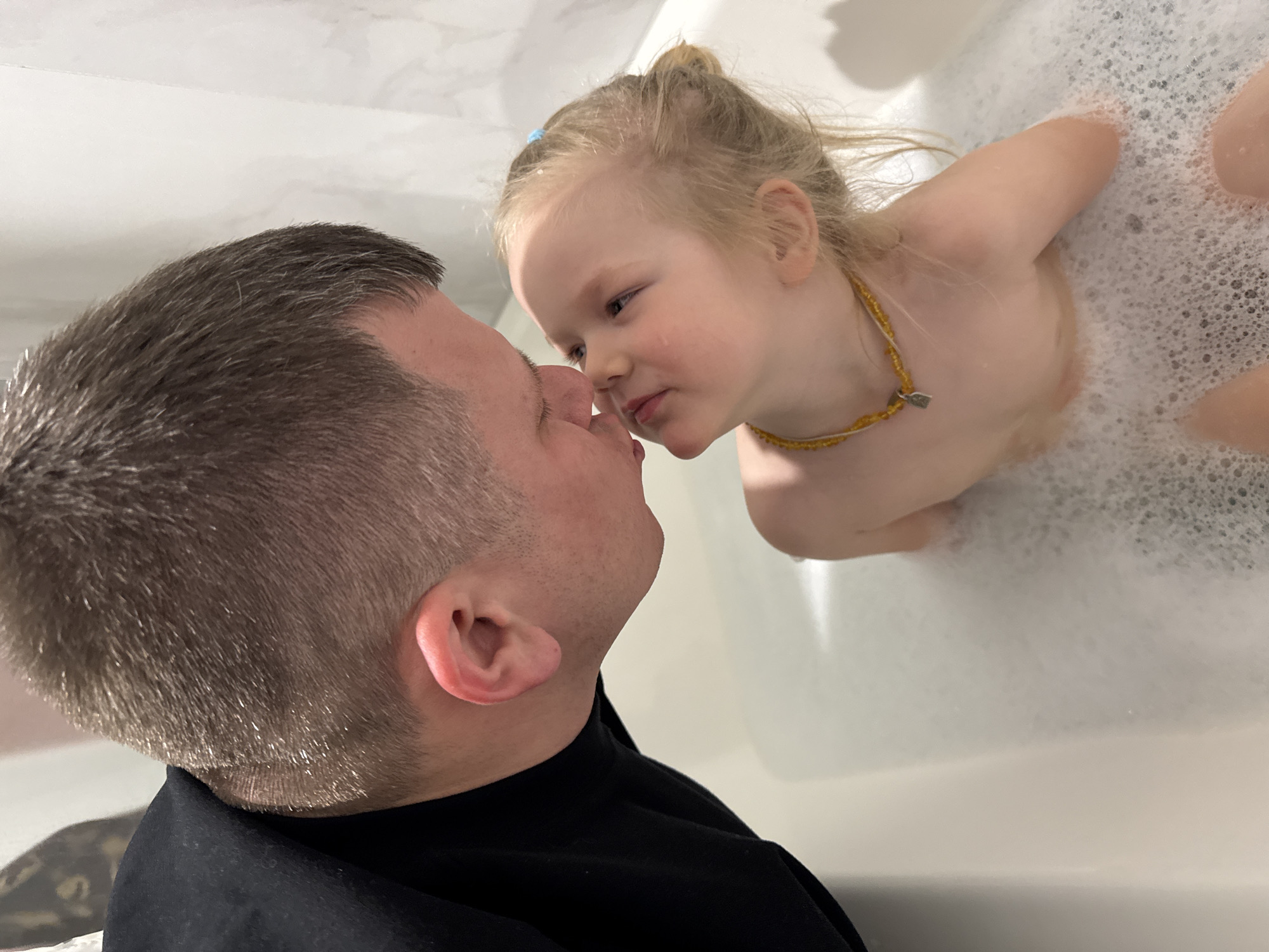 A man and a young girl are interacting in a bathroom, likely in a domestic setting. The man, positioned on the left, has short, graying hair, a fair complexion, and is wearing a black shirt. He appears to be leaning forward, his face close to the girl's. The girl, on the right, is in a bathtub filled with bubbles. She has blonde hair pulled back, and is wearing a beaded necklace. Their faces are close, almost touching. The environment appears to be a standard bathroom with white walls and fixtures. No identifiable elements suggest the location in Ферма, Russia. The interaction between the man and girl appears affectionate.