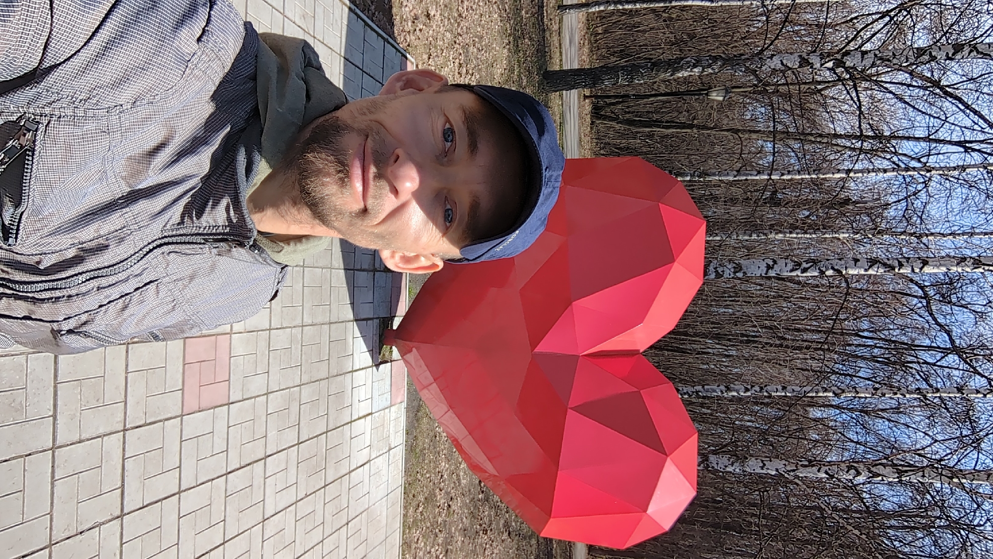 A man with a light beard, blue eyes, and a slight smile looks directly at the camera in this outdoor portrait. He is wearing a dark blue baseball cap with faint white text, and a light blue and grey checkered jacket over a greenish hooded garment. He stands on light grey tiled pavement, which features occasional reddish-pink accent tiles.
To his right, a prominent, large red geometric heart sculpture is visible. The sculpture is faceted, made of many flat panels, giving it a crystalline appearance. In the background, a clear bright blue sky stretches above a line of trees with bare, dark branches and characteristic white bark, identifiable as birch trees. The ground beneath the trees is covered in dry leaves or sparse vegetation.
The image appears to be a selfie or a close-up photograph taken on a sunny day, indicated by the strong lighting and shadows. The bare trees suggest either late autumn or early spring. The scene captures a moment of engagement between the man and the striking heart sculpture. The location context is Ulyanovsk, Russia.
