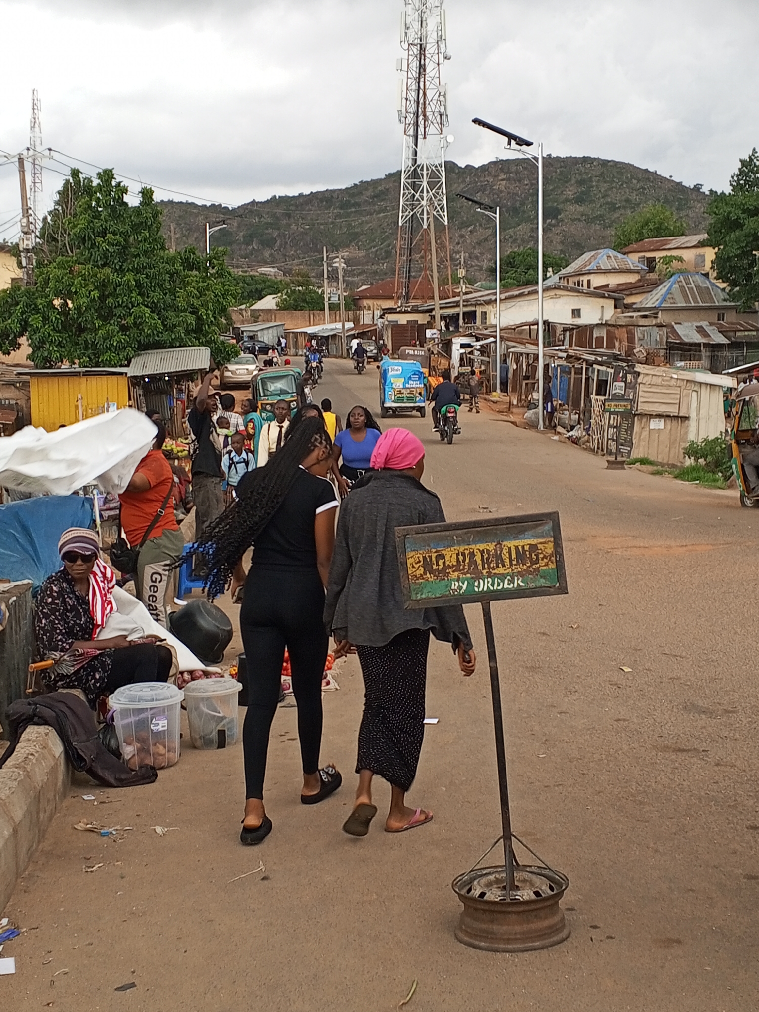 In Gwong, Nigeria, a street scene shows pedestrians and vehicular traffic along a road. Two women walk on the right side of the road, one in black attire and the other in a grey jacket and pink headscarf, alongside a rusty "NO PARKING BY ORDER" sign anchored by a wheel rim. A woman sits on the curb, selling items displayed in plastic containers. Various people are gathered along the street, with some interacting and others engaged in commerce. Vehicles, including motorcycles and auto-rickshaws, move along the street, adding to the urban activity. Buildings line the street, with a hill visible in the background. A tall communication tower rises above the buildings, partially obscured by trees. The sky is overcast.