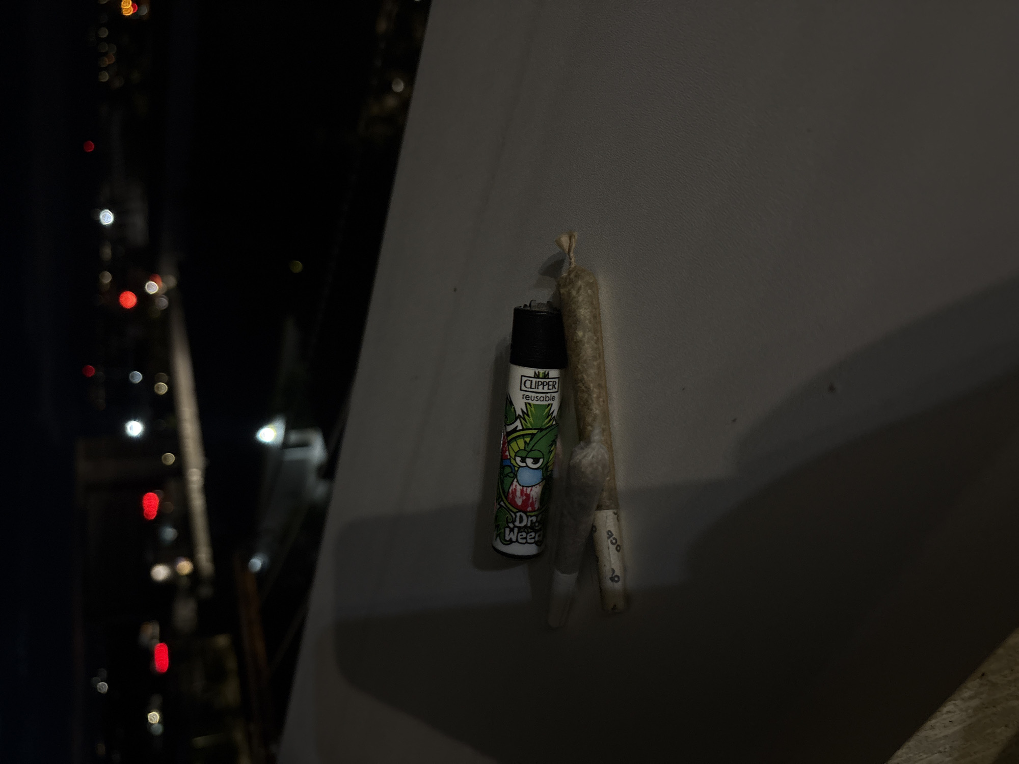 The image depicts a Clipper lighter with a "Dr. Weed" design, and two cannabis joints on a gray surface. The background suggests an urban setting at night, with blurred lights resembling city traffic and buildings. The location is potentially Sofia, Bulgaria, as specified in the instructions.