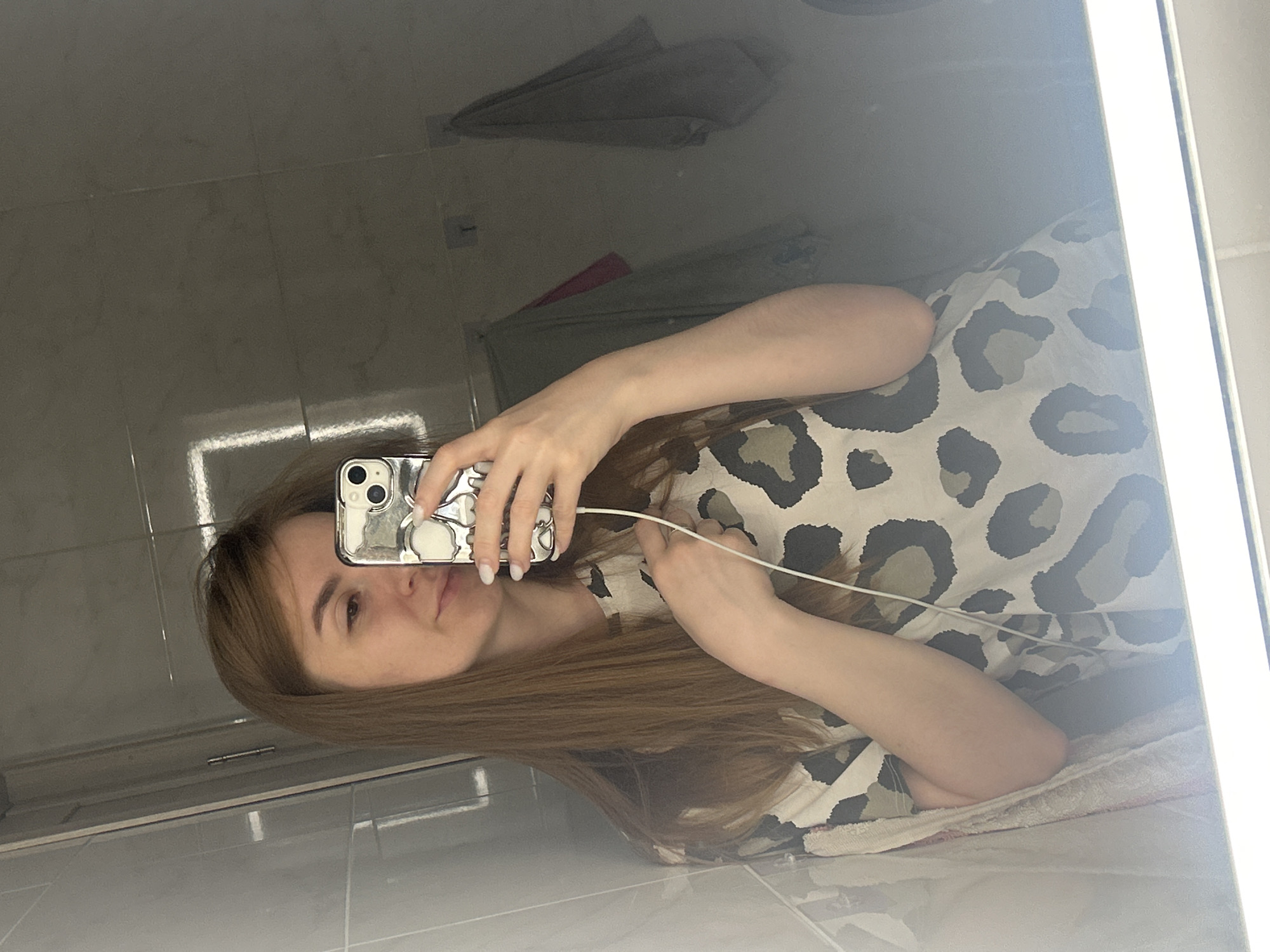 A young Caucasian woman with long brown hair is taking a selfie in a bathroom. She is wearing a light-colored t-shirt with a leopard print pattern. Her right arm is extended, holding a smartphone with a silver skull-patterned case. A white cord is visible extending from the phone. The background is a tiled bathroom wall with a large mirror reflecting the scene. Two towels are hanging on the wall behind her. The lighting suggests an artificial light source above the mirror. The location is the bathroom of a residential dwelling in Нижние Муллы, Russia.