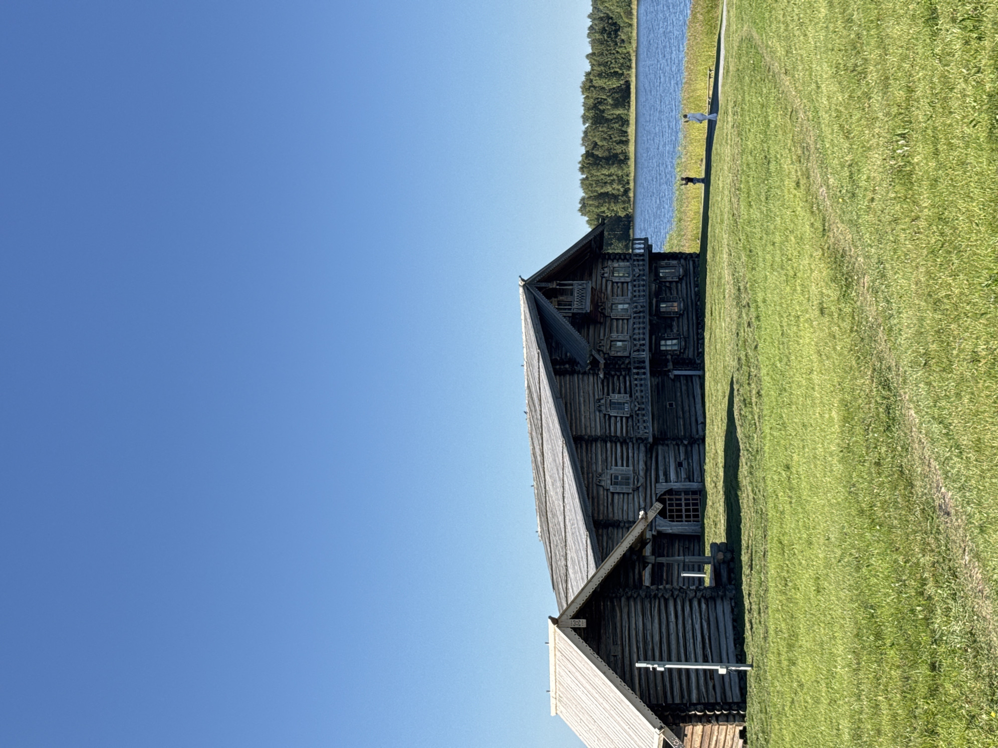 The image shows a bright, sunny day with a clear blue sky over a grassy field. In the foreground, a well-maintained path winds through the vibrant green grass. To the left, two traditional wooden houses, characteristic of Russian architecture, stand side-by-side. The larger house is multi-story with intricate wooden detailing and balconies, while the smaller one is a simpler structure. Both are constructed from horizontal logs and have wooden roofs.
In the middle ground, a calm blue lake stretches across the scene, bordered by a dense line of green trees forming a treeline on the far shore. The water reflects the clear sky and the surrounding greenery.
Further to the right, near the lake's edge, two figures are visible. One appears to be a child in dark clothing, facing away from the viewer, and another figure in white clothing, also facing away, is standing to their right. A bench is situated behind the figure in white.
The scene depicts an idyllic outdoor setting in Boyarshchina, Russia, with the rustic charm of the wooden buildings set against the natural beauty of the lake and forest.