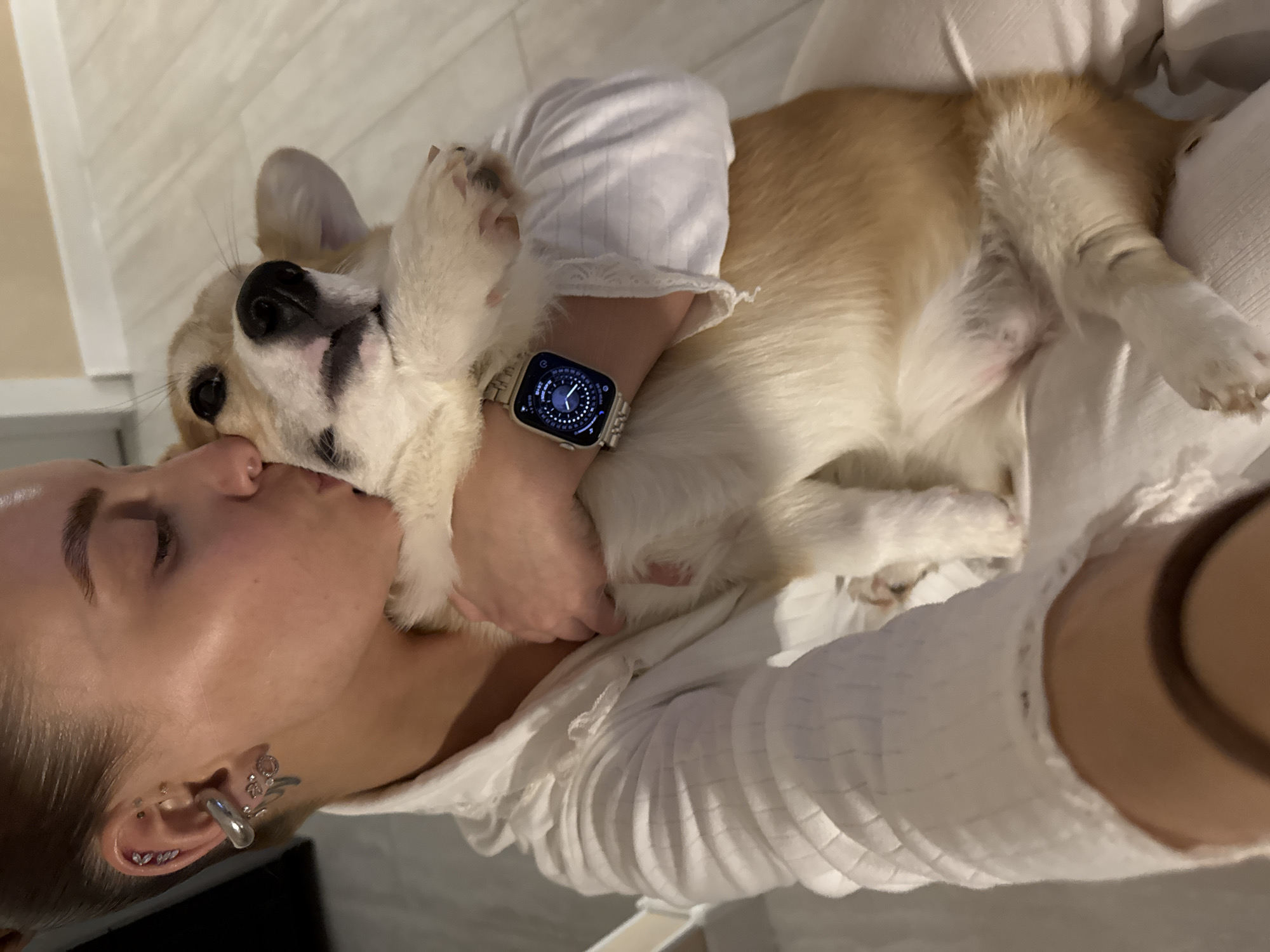The image captures a heartwarming moment indoors, likely in Chaikovsky, Russia, showing a young woman affectionately kissing her Corgi dog. The woman has light hair tied back and multiple ear piercings, including a large silver cuff, studs, and smaller hoops. She is wearing a white or light-colored top with a subtle quilted or textured pattern. Her eyes are closed as she leans in to kiss the dog's head, conveying deep affection and comfort.
The Corgi, with its distinctive light brown and white fur, is nestled close to the woman, appearing relaxed and content. It is wearing a white, short-sleeved garment on its front torso, possibly pajamas or a casual shirt. A silver smartwatch with a complex digital face, featuring concentric circles and indicators for time or data, is prominently displayed on the woman's left wrist, resting against the dog's fur. Close-up views confirm the dog is male, with visible testicles. The background is softly lit and consists of light-toned surfaces, suggesting a cozy home environment. The scene exudes warmth, intimacy, and the strong bond between a pet owner and her dog. No specific text is clearly legible on the smartwatch or within the image.