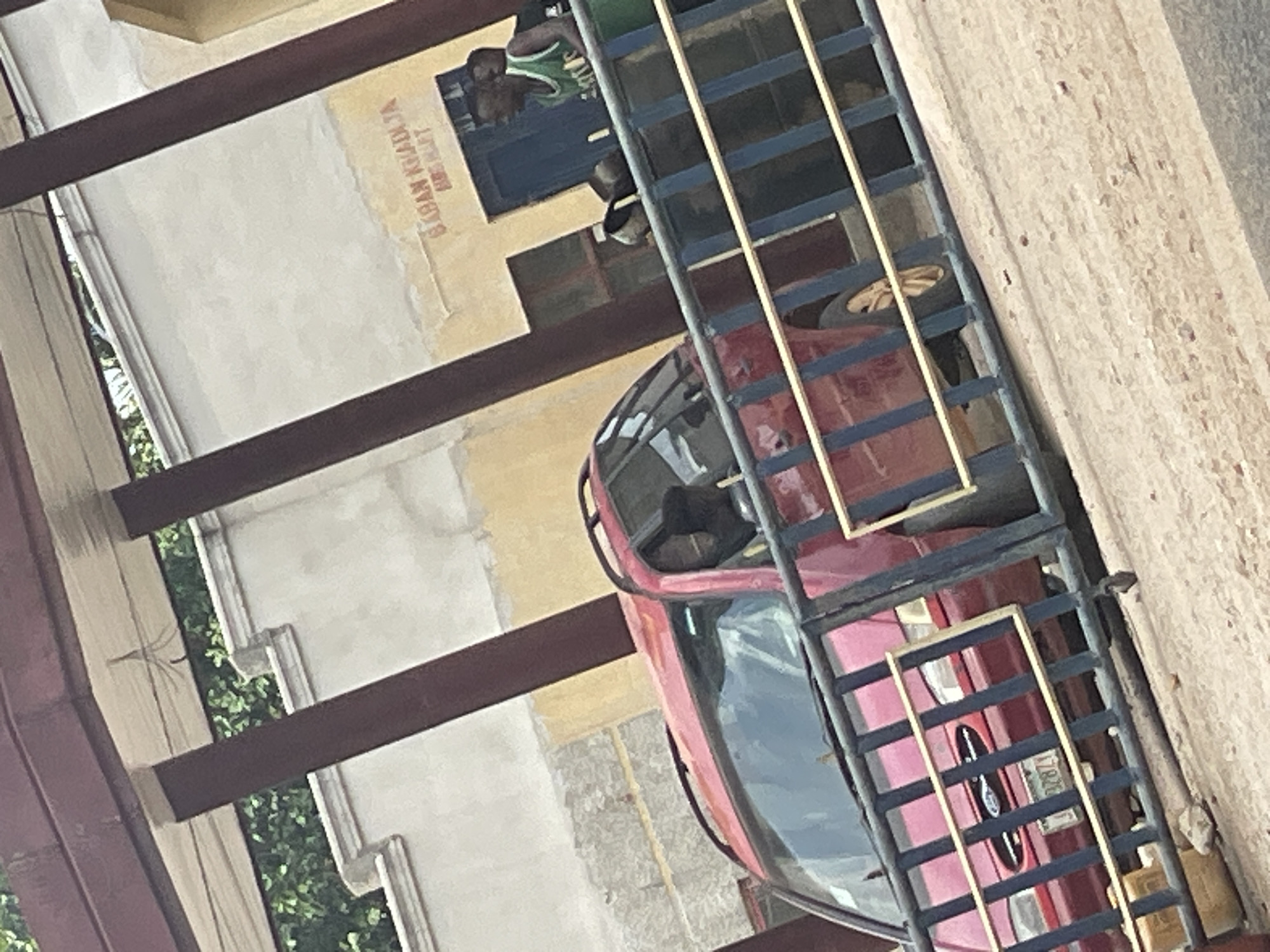 The image, when viewed upright, captures an outdoor scene under bright daylight, likely in Mararaba, Nigeria. A red car, possibly a sedan, is prominently featured, partially obscured by a dark metal structure in the foreground. This structure appears to be a gate, fence, or a robust external cage, characterized by dark horizontal and vertical bars accented with lighter, possibly yellow or beige, elements.
To the left of the car, a building with light-colored walls, featuring shades of yellow and off-white or grey, is visible. A person wearing a green and white patterned tank top can be seen looking out from a dark blue-framed window opening on the building. Another individual, largely obscured by the metal structure, is situated near the red car, dressed in dark clothing.
Green foliage is visible in the background, behind some dark brown horizontal structural beams attached to the building. The ground to the right of the car appears to be a light-colored paved or dirt surface.
Faint red text is visible on the building wall, positioned above the blue window. The text clearly reads "GABAN KHADIJA" with additional, partially obscured text below it that appears to be "MRS N..." possibly "MRS NADIYA" or similar. The clear lighting indicates it is daytime.