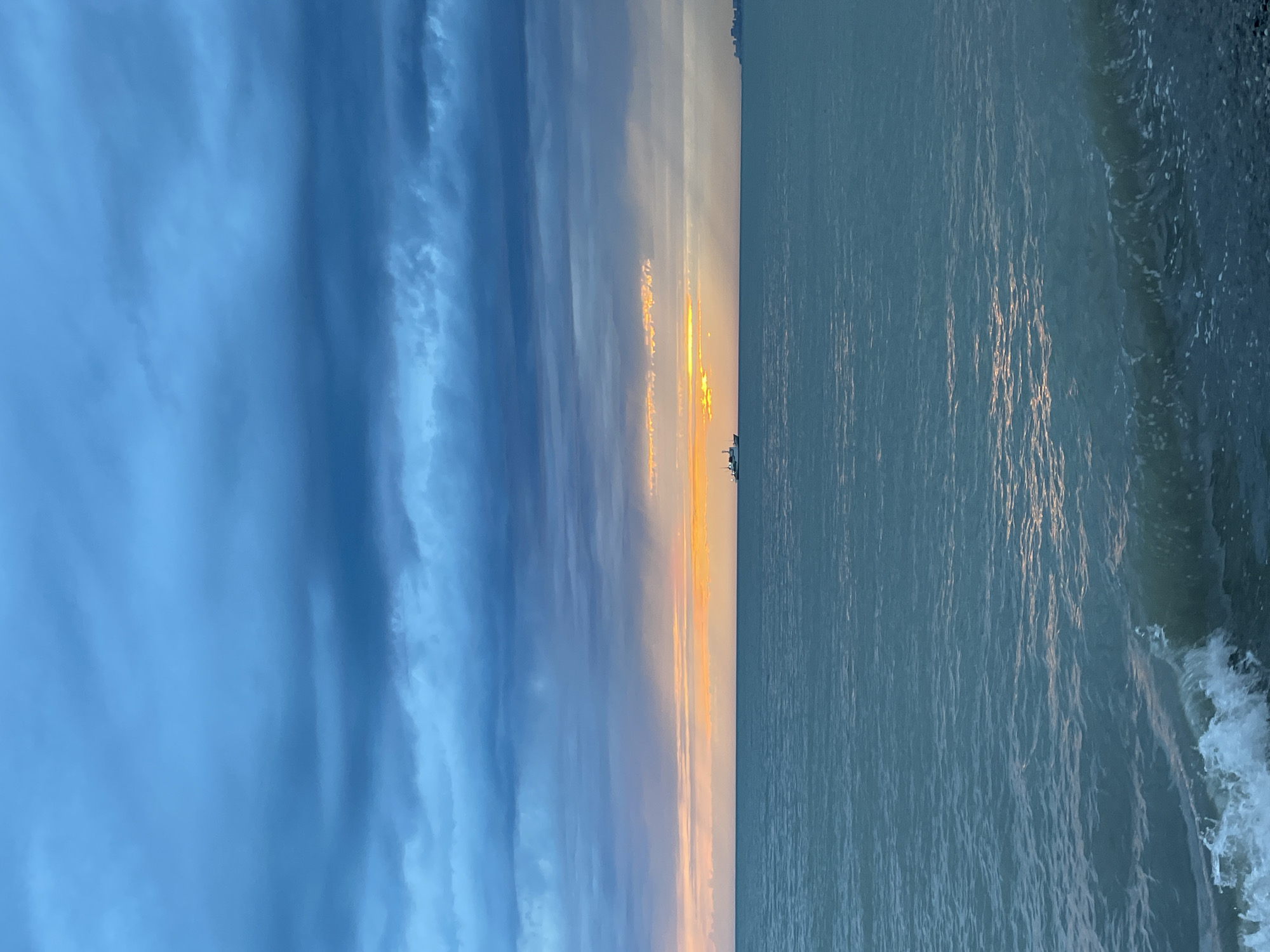 The image depicts a shoreline at sunset. The sky on the left side of the image is a gradient of dark to light blue with textured clouds. A thin horizontal band of orange and yellow light separates the sky from the water. The water, on the right side, is a pale blue with textured ripples. The shore, along the right edge, shows the movement of waves. The horizon is visible in the center of the image, with a couple of ships on the horizon. There appear to be some buildings on the horizon as well. The location is likely in Sochi, Russia based on the buildings on the horizon, although not definitely. The time is evening based on the lighting.
