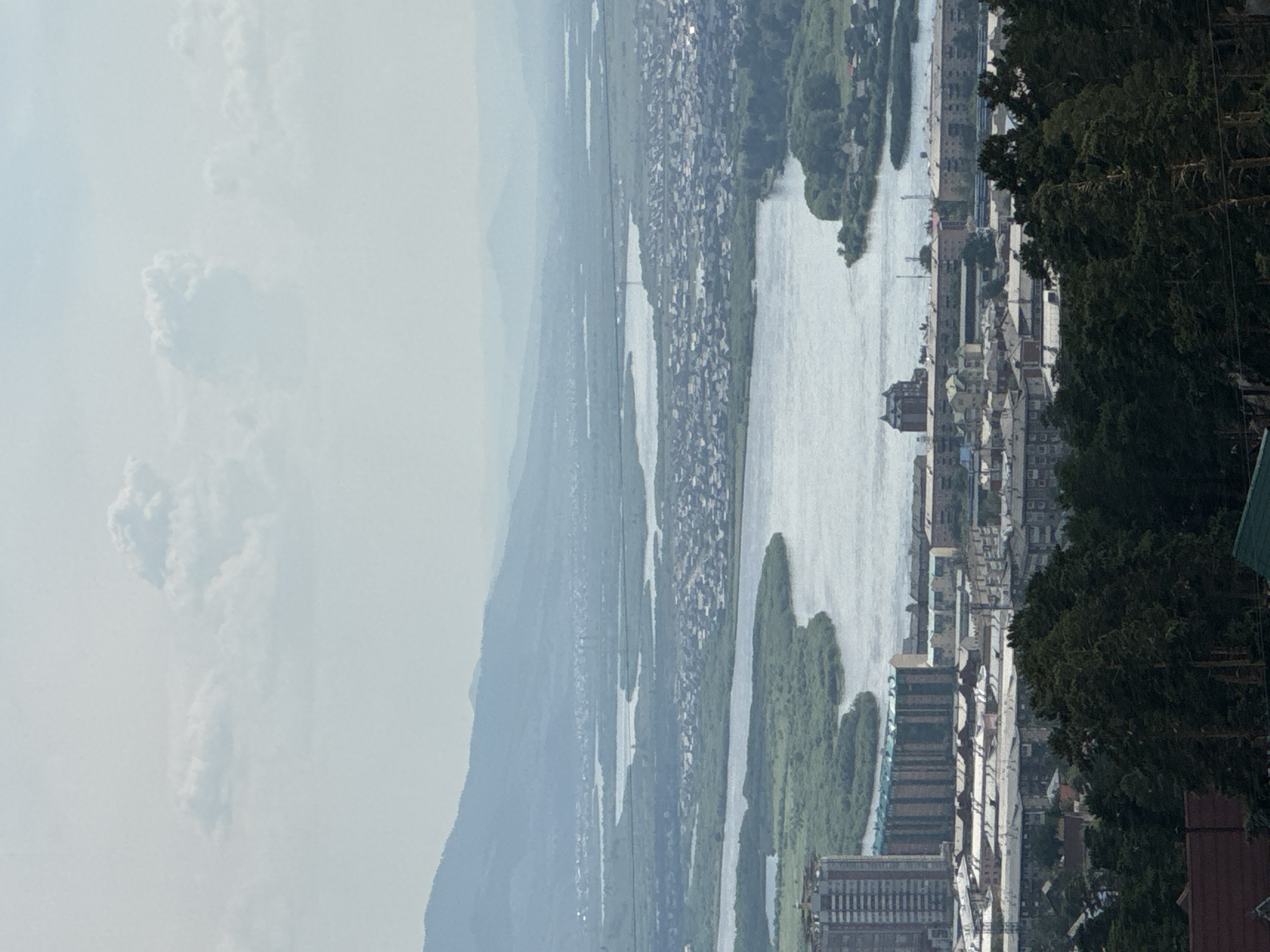 The image displays a panoramic view of the city of Ulan-Ude, Russia, on a hazy day. In the foreground, a dense forest of pine trees is visible, with some rooftops of houses peeking through. Beyond the trees, the cityscape unfolds with a mix of residential buildings and a prominent tall building with a striped facade. A wide river, likely the Uda River, snakes through the urban landscape, with islands of lush green vegetation along its banks. In the background, a vast expanse of plains stretches towards a range of misty blue mountains. The sky is overcast with scattered clouds.