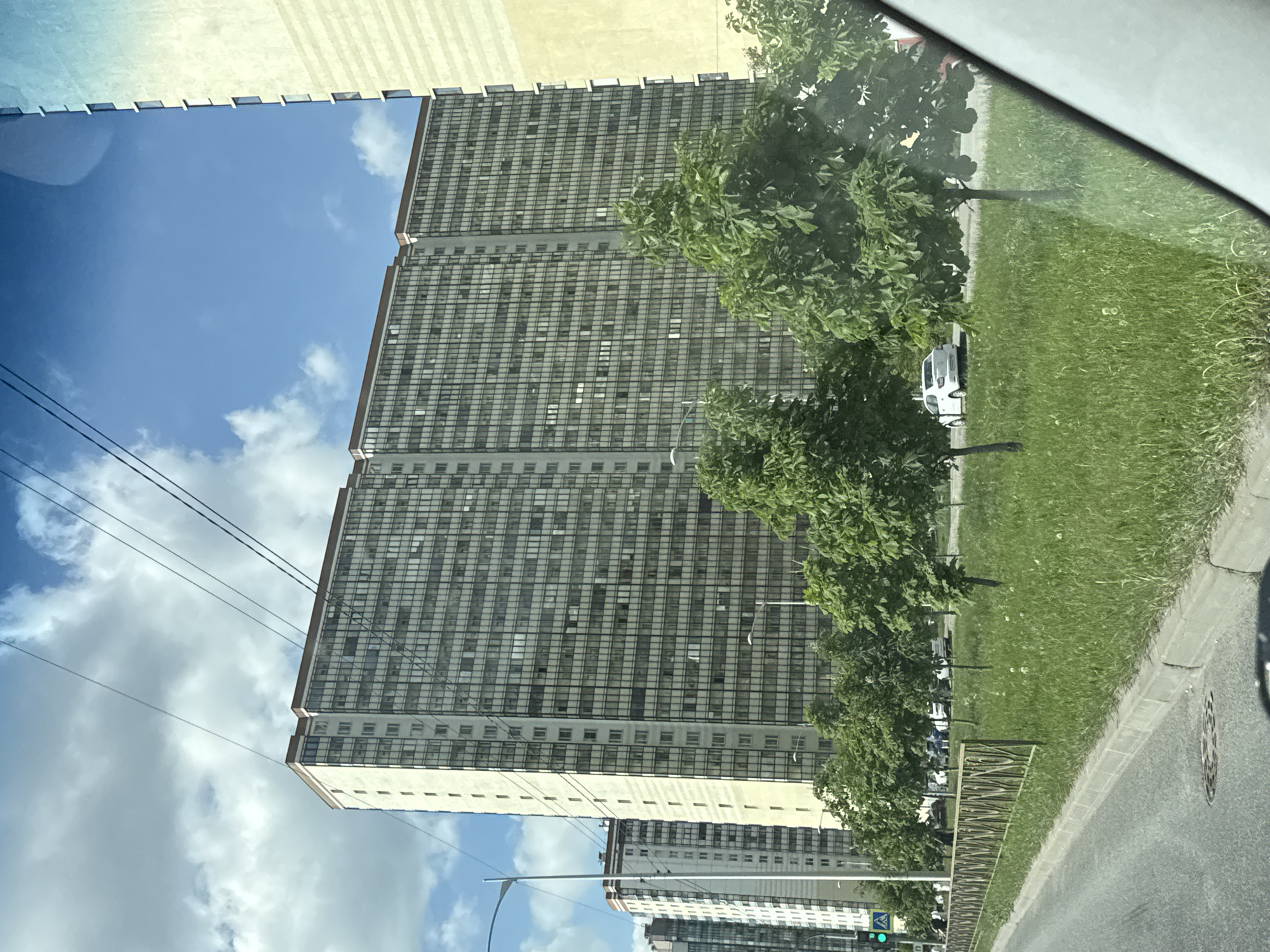 The image, originally rotated 90 degrees clockwise, captures an urban or suburban street scene on a bright, cloudy day, seemingly from inside a vehicle. On the left, two prominent, tall multi-story buildings dominate the skyline, featuring a grid-like facade of numerous uniform windows, suggesting residential or office use. A third, similar building is visible further down the street. The buildings stand against a blue sky dotted with scattered white clouds.
To the right, a wide strip of vibrant green grass is punctuated by several lush, green deciduous trees. A paved road with a clear curb runs alongside the grassy area. A white car is partially visible behind trees, and another white car can be seen further down the road. Infrastructure includes dark power lines stretching across the upper left, a street light pole in the lower right, and a circular manhole cover on the road. A low, metal fence borders a section of the grassy area. The diagonal lines of a car's interior or window frame are visible along the top and far right edges, indicating the vantage point. It is daytime with ample natural light. No people or specific activities are discernible. There is no visible text. The architectural style and urban setting are consistent with areas in Russia, matching the provided context of Poroshkino.