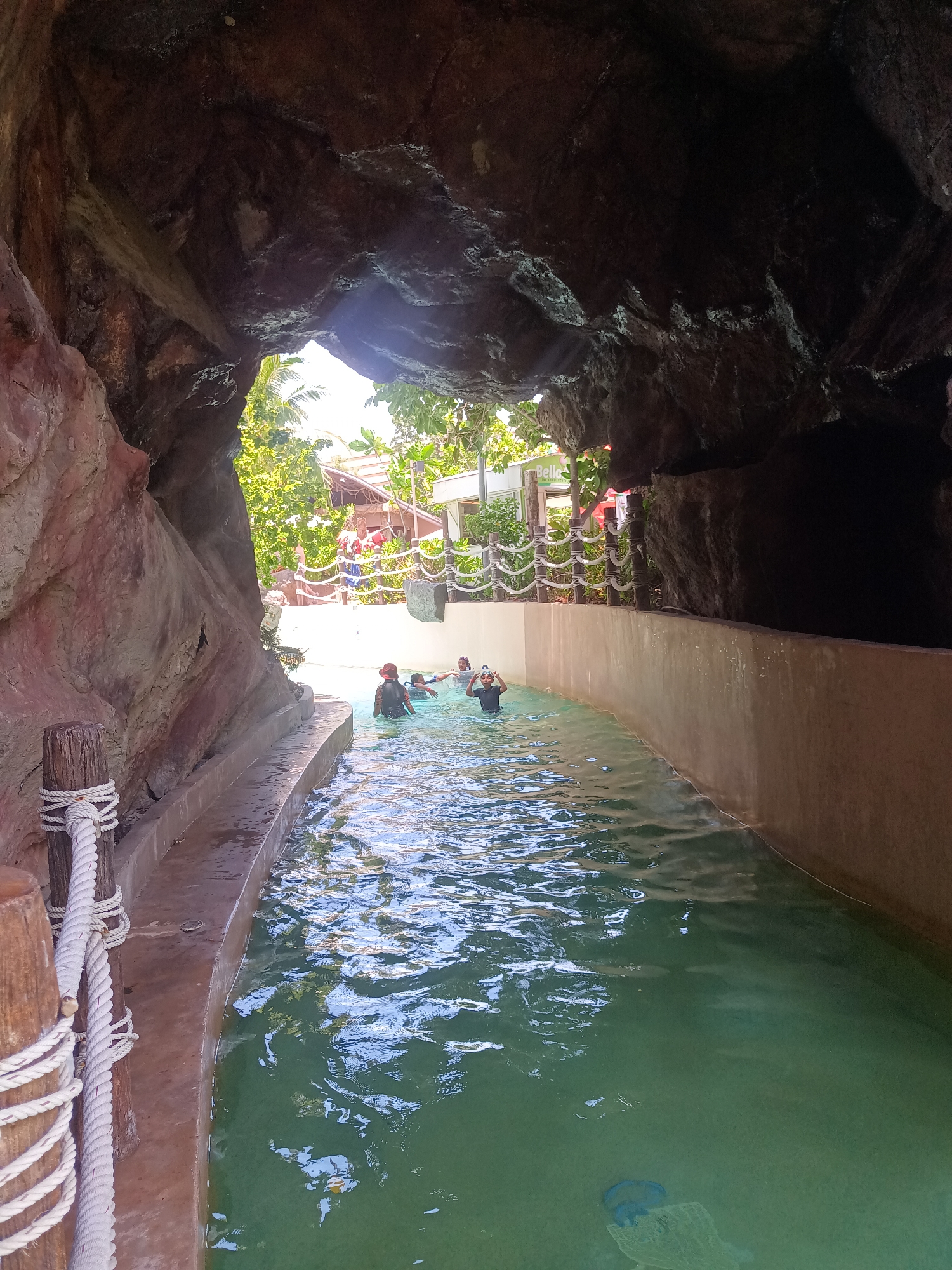 The image depicts an outdoor water park attraction in Hua Hin City Municipality, Thailand. The perspective is from within a dark, cave-like structure overlooking a shallow, artificial river channel. In the channel, several people are visible, some wearing swimsuits and water shoes, enjoying the attraction. Outside the cave opening, there is a fence and some buildings and trees. There are also some posts on the left with thick white ropes wrapped around them. The water is green and some objects can be seen at the bottom of the channel.