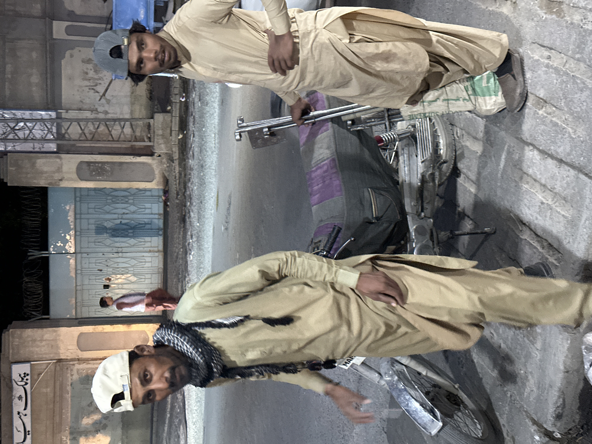 Two men wearing tan clothing are standing next to a motorcycle in a city location. The man on the left is wearing a white cap and a patterned scarf. The man on the right is wearing a gray baseball cap. The motorcycle is parked on the street, with a bag on the back. Buildings are visible in the background.