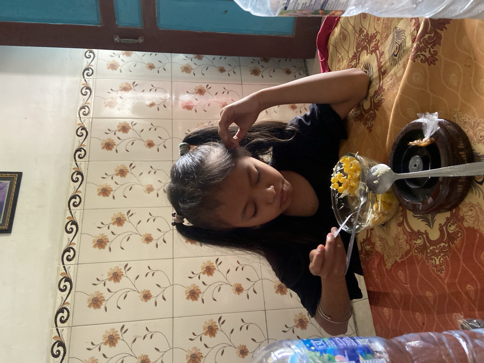 A young girl with pigtails is eating from a bowl of corn and what appears to be cheese. She is sitting at a table with a patterned tablecloth, in what seems to be a home in Baron, Indonesia. There is a bottle of water on the left side of the image and another bottle of water on the right side. The wall behind the girl is tiled with a floral pattern. A doorframe is visible on the right side of the image.