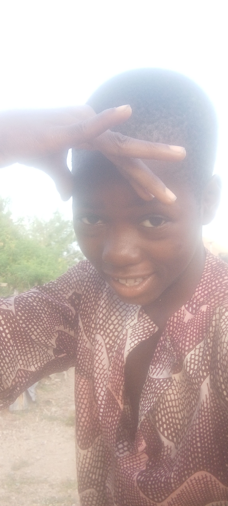 A young person with dark skin poses for a photo. Their left hand is raised to their forehead, fingers extended and slightly apart. They are wearing a patterned shirt with a reddish-brown base and light-colored details. The background is blurred, but green foliage suggests an outdoor location. The image was taken in Adankolo, Nigeria.