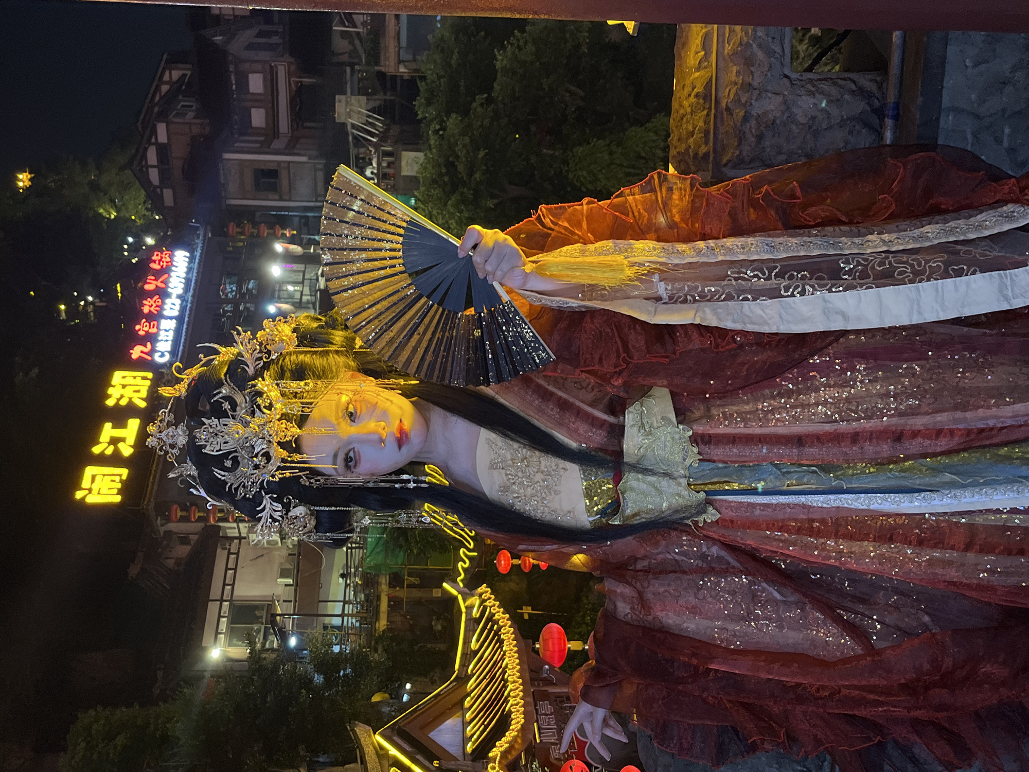 A young woman in traditional Chinese attire, including an elaborate headdress and a flowing, multi-layered red and gold gown adorned with sequins, stands outdoors at night. She holds a black and gold fan in her right hand, positioned in front of her face. Her left arm is extended slightly, with her hand open. The background features buildings with illuminated signage and traditional architectural elements, suggesting a location in a historic or culturally themed district. The yellow neon sign in the background reads "阎江湖," which translates to "Yan Jiang Hu," a name that could be associated with a restaurant or establishment. Based on the architectural style and the signage, this scene is likely located in a place like Hongyadong in the Yuzhong District of Chongqing, China, a popular tourist area known for its traditional stilt houses, night markets, and vibrant cityscape, especially at night. The lighting in the image is predominantly from artificial sources, creating a dramatic and atmospheric effect.