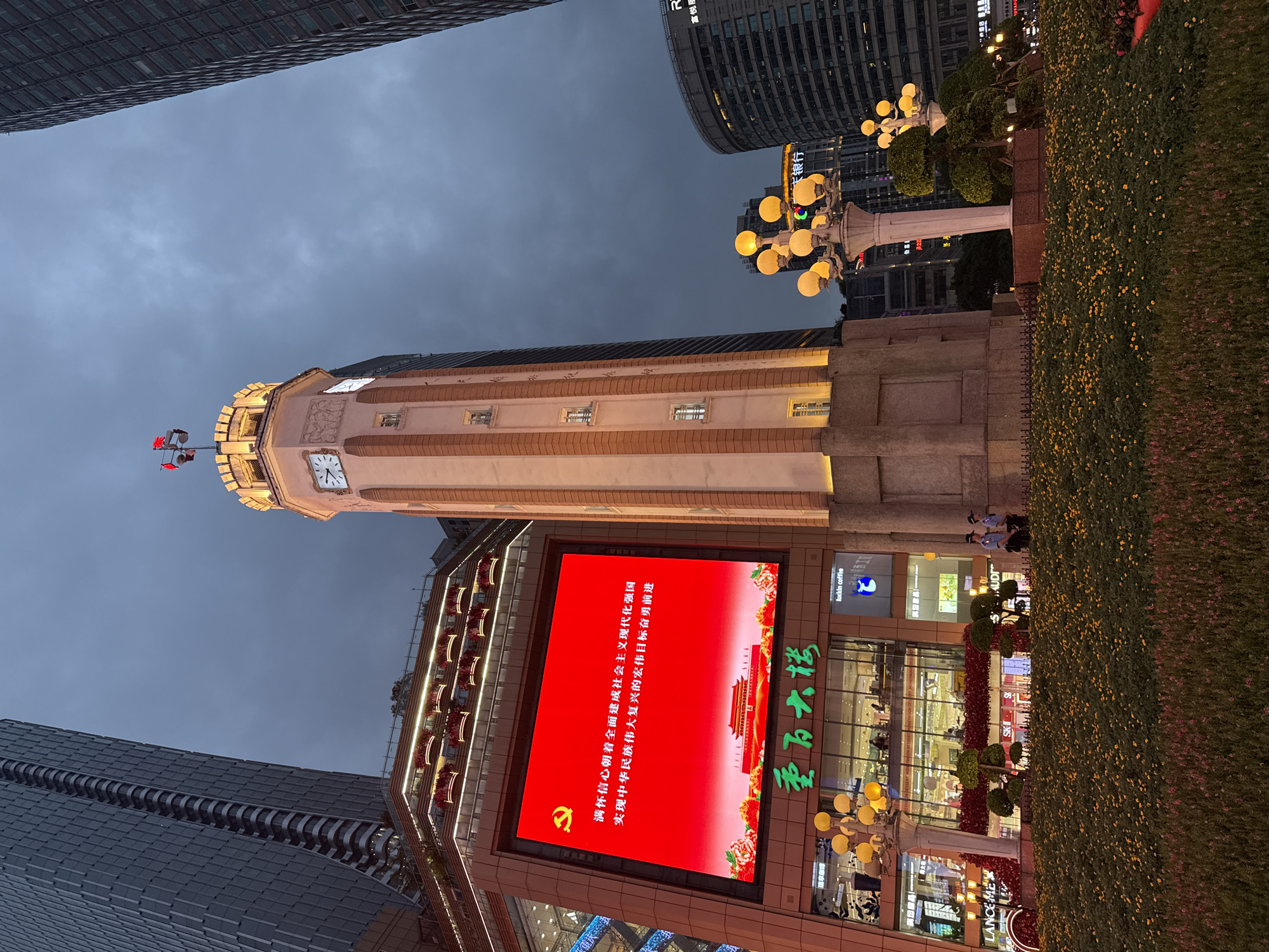 In the Yuzhong District of China, a tall clock tower stands prominently in a city square at dusk. The tower, with its light-colored facade and red brick accents, features illuminated clock faces and decorative carvings. It is topped with a small structure flying two red flags. Adjacent to the tower is a large, brightly lit red LED screen displaying a message in Chinese characters along with an image of a historical building. Below the screen, the facade of a multi-story building, likely a commercial establishment, is visible with glowing signs and windows. In the foreground, a meticulously maintained flower bed with numerous small yellow flowers lines a paved area. Two uniformed individuals, possibly security personnel, stand near the flower bed. Several ornate lampposts with multiple yellow lights illuminate the scene. The background is dominated by tall, modern skyscrapers under a cloudy, twilight sky.