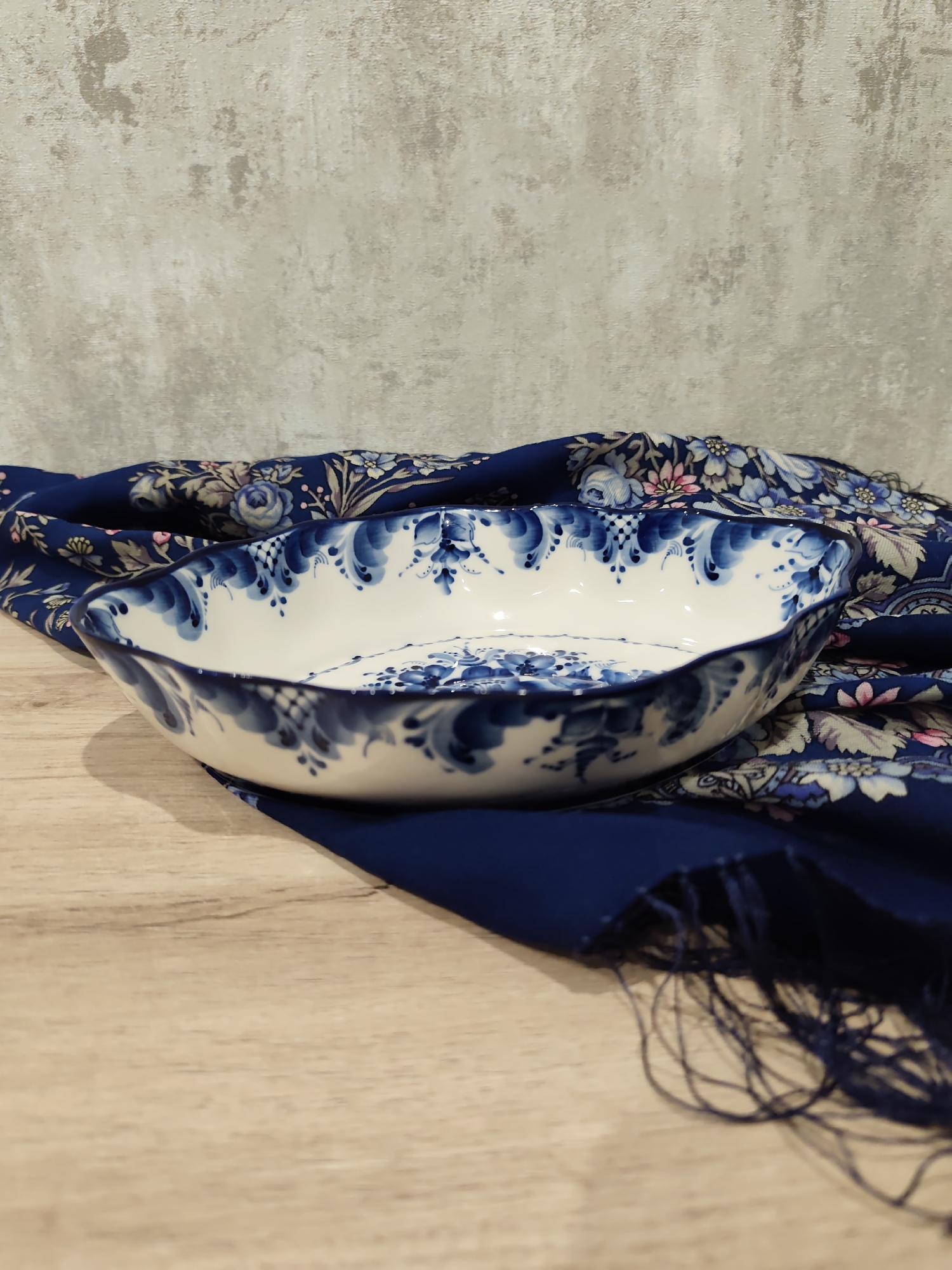 A close-up, eye-level shot shows a white oval bowl with blue Gzhel painting. The bowl rests on a navy blue scarf with a floral pattern. The scarf has tasseled edges and is draped over a light brown wooden surface, which is likely a table. In the background, a grey, textured wall serves as the backdrop. The scene appears to be indoors, possibly in a home or a shop, and there are no people present. The lighting is soft, creating subtle shadows. The overall impression is one of decorative artistry. The location is not specified in the image, so it cannot be determined if it is in Sochi, Russia.