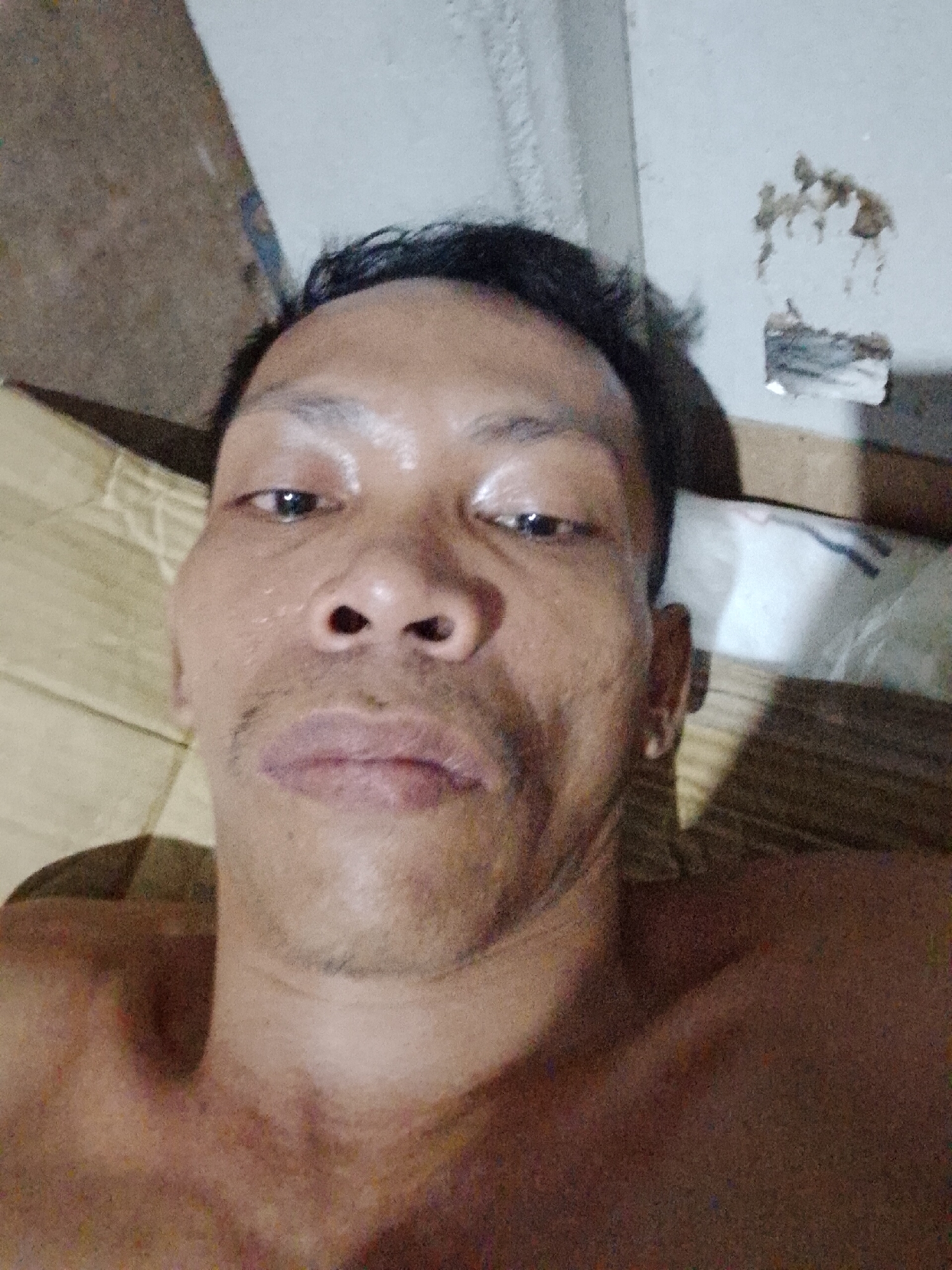 A close-up, low-angle selfie of a shirtless man with dark hair, appearing slightly sweaty. He has an Asian facial structure with a light mustache, and is looking directly at the camera. The background is out of focus but shows a section of a brown corrugated cardboard surface, what appears to be a white bag and a wall with some discoloration and an electrical outlet. The location is unknown. There is no information to confirm the location is in Bukit Ketok, Indonesia.