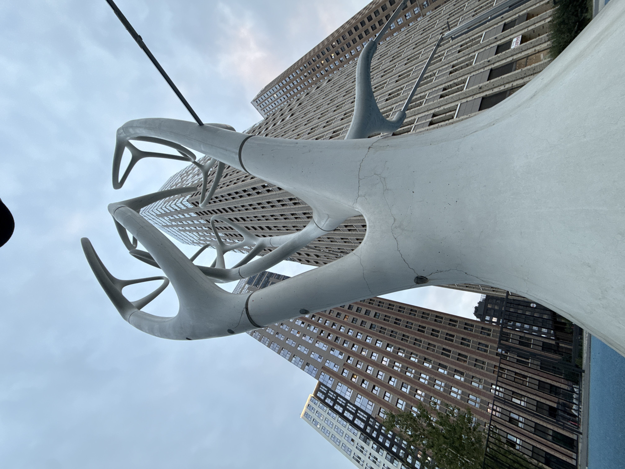 This is a low-angle, full shot of a large, grey, abstract sculpture resembling a tree or branching structure. The sculpture is in the foreground, dominating the lower two-thirds of the image. It has a thick, curved base and branches out into thinner, rounded arms with hollow, triangular ends. The sculpture appears to be made of concrete or a similar material and shows some cracks and imperfections.
Behind the sculpture, two tall, modern apartment buildings are visible. The building on the left is lighter in color with red brick accents, while the building on the right is a darker, uniform grey with many small, regularly spaced windows. The buildings are tilted due to the low-angle perspective. The sky is overcast and a pale grey. A thin, dark cable runs diagonally across the upper right portion of the image, intersecting the sculpture. In the lower left, a black metal fence and a section of blue ground or surface are visible. A few small trees are also present near the fence line. The scene appears to be in an urban environment, possibly a park or public space.