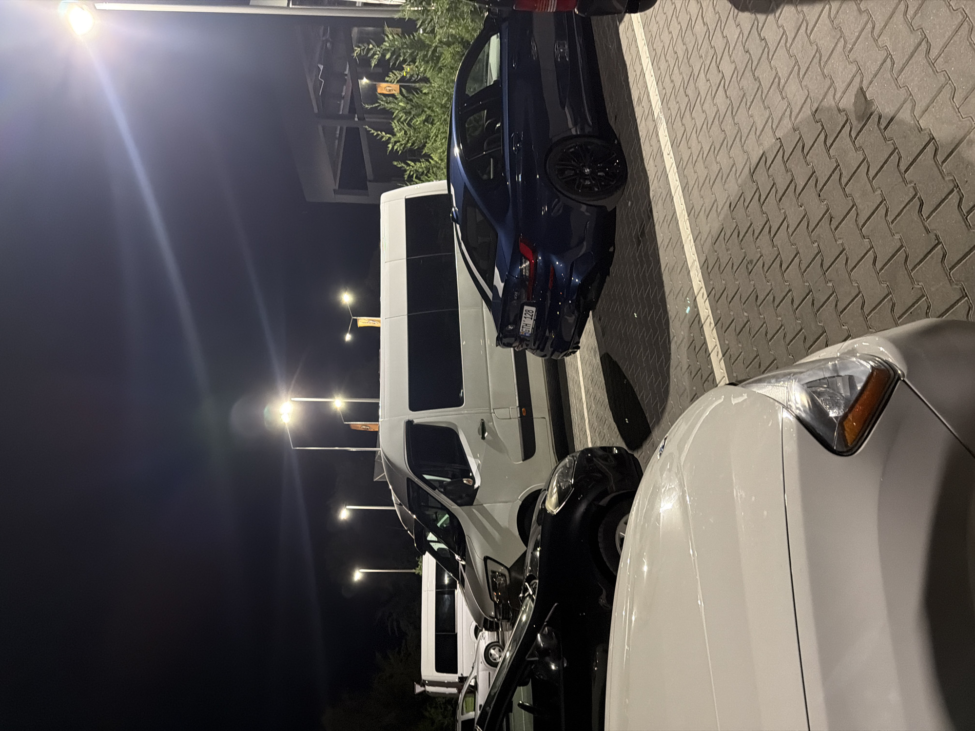 At night, on a paved lot in Băcioi, Moldova, a dark blue BMW sedan is parked next to a white van. In the foreground, the hood of a white car is visible. The scene is illuminated by streetlights, casting shadows.