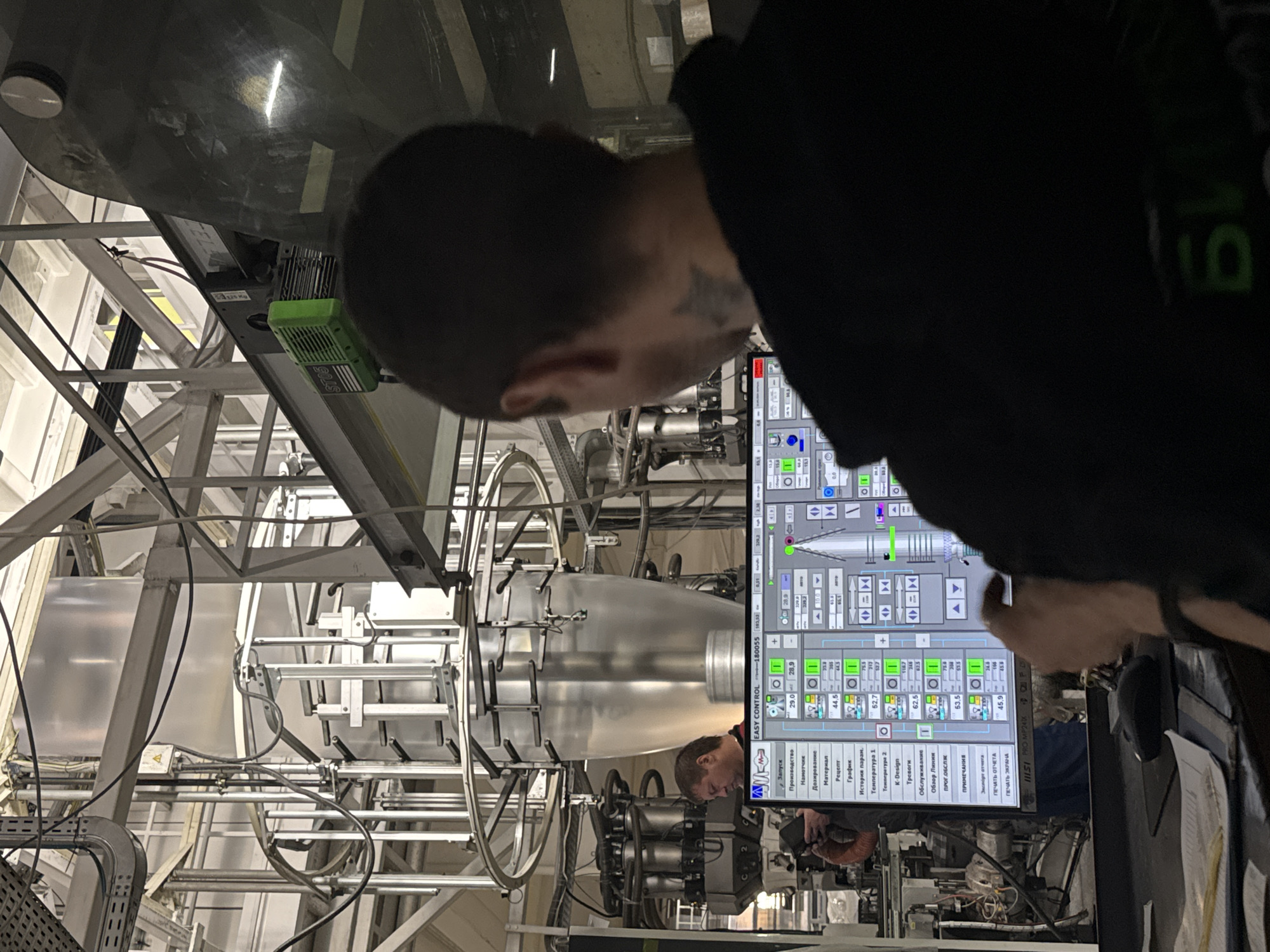 The image shows an industrial setting, likely a manufacturing facility. A person with short dark hair, visible ear, and a tattoo on their head is viewing a computer screen showing "EASY CONTROL" software with Russian text, likely operating a large machine with various silver pipes and components. Based on the OCR'd text, which contains the software's language, this facility is likely in Perm, Russia. The individual is wearing a dark-colored top with a green logo on the sleeve. The computer screen displays various controls and data points related to production processes. The environment includes industrial equipment, metal structures, and overhead lighting.