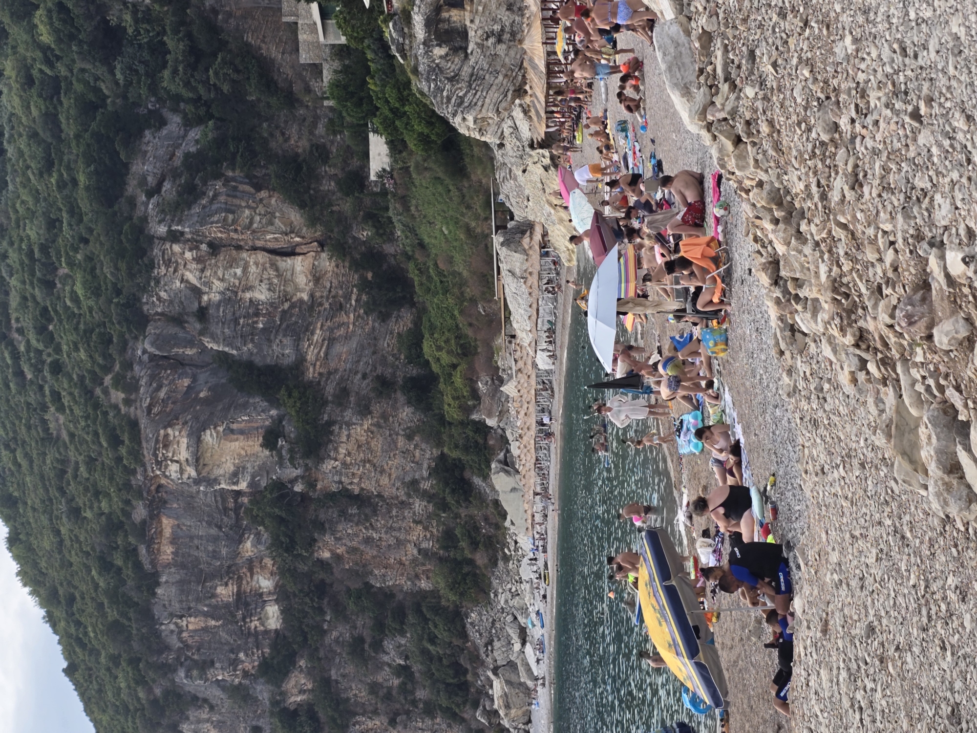 The image depicts a crowded pebble beach in Perazića Do, Montenegro, situated at the base of a steep, vegetated cliff face. Numerous individuals are engaged in various beach activities. In the foreground, several people are relaxing on the pebbled shore, some lying on towels or beach chairs, while others sit or stand. One person in a wetsuit appears to be interacting with another individual lying on the ground. Further back, closer to the water's edge, a yellow and blue inflatable boat is partially visible, with people in and around the water. The sea is calm and appears to be a popular area for swimming and recreation. A line of beach umbrellas and lounge chairs is set up further along the beach, providing shade for sunbathers. The rocky cliffs rise dramatically behind the beach, covered in dense green foliage, with some structures visible on the upper slopes. The overall scene is one of a busy, popular summer vacation spot.
