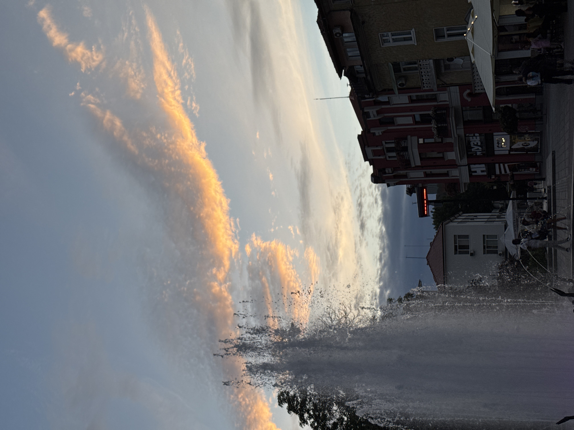 This is a photograph taken in Plovdiv, Bulgaria, likely in the early evening, as indicated by the soft, warm light illuminating the sky and the clouds. The scene captures a public space with a prominent fountain in the foreground, its water spraying upwards in a dynamic display. In the background, several buildings are visible, displaying a mix of architectural styles. The building on the right is painted yellow with white window frames and balconies. Adjacent to it is a taller building with a red facade, featuring decorative white elements and multiple balconies. Further to the left, a white building with visible windows and what appears to be an air conditioning unit is present. The sky above is a canvas of blues and grays, with streaks of orange and pink clouds suggesting a sunset. Several people are present in the scene, some walking near the fountain, others appearing to be seated at outdoor tables under umbrellas, suggesting a cafe or restaurant setting. A sign with Cyrillic lettering, potentially indicating a shop or establishment, is visible on the red building. The overall atmosphere appears relaxed and urban.