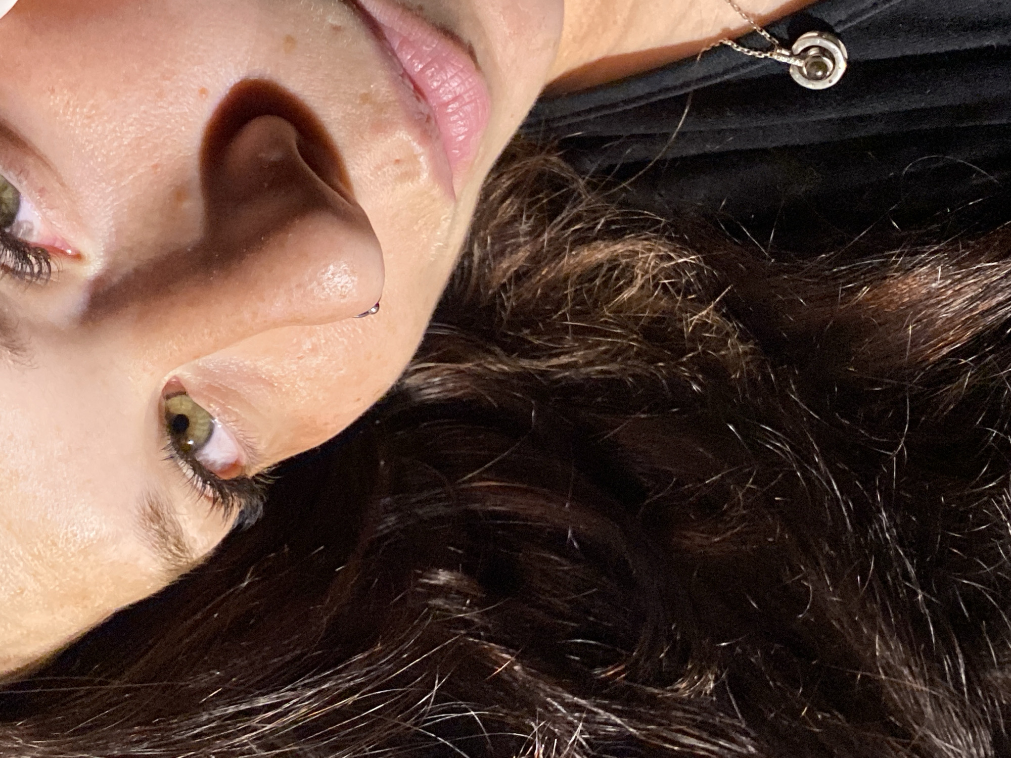 The image is a close-up, low-angle, headshot of a person. The subject is wearing a black top and a necklace with a circular pendant. The person has dark brown hair, green eyes, and a nose piercing. The lighting suggests the image was taken indoors with artificial light. The scene does not contain any elements that would indicate a specific location in Chaikovsky, Russia.