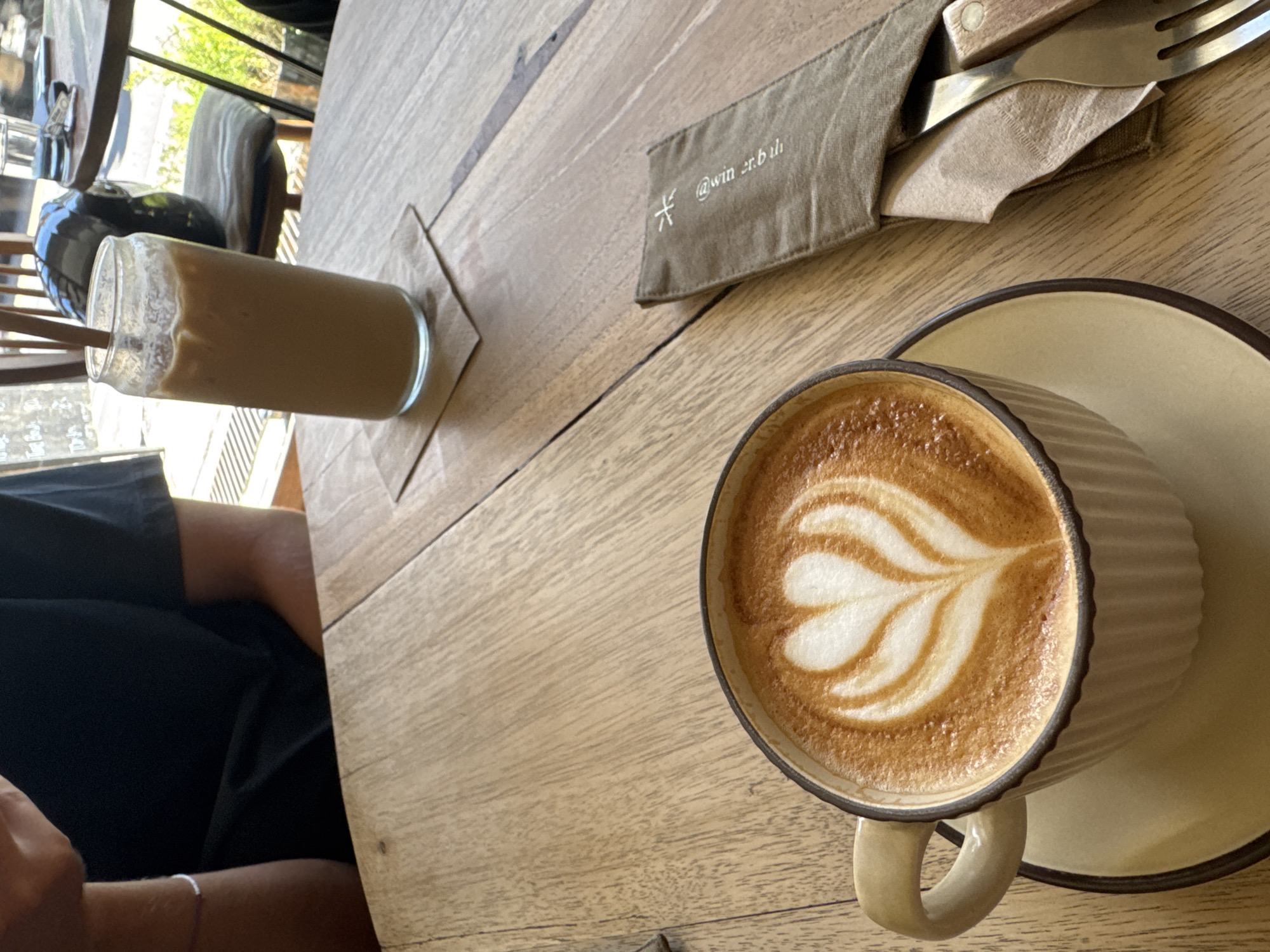 A high-angle shot shows a table with beverages and utensils.
A glass cylinder containing a light brown liquid rests on a square coaster. A khaki fabric napkin, embroidered with a symbol and "@win.er.buli", lies near a wrapped utensil set. A coffee cup, with latte art, sits on a matching saucer. A person is partially visible, wearing a black shirt and a thin bracelet.
The location is Unknown, Unknown.