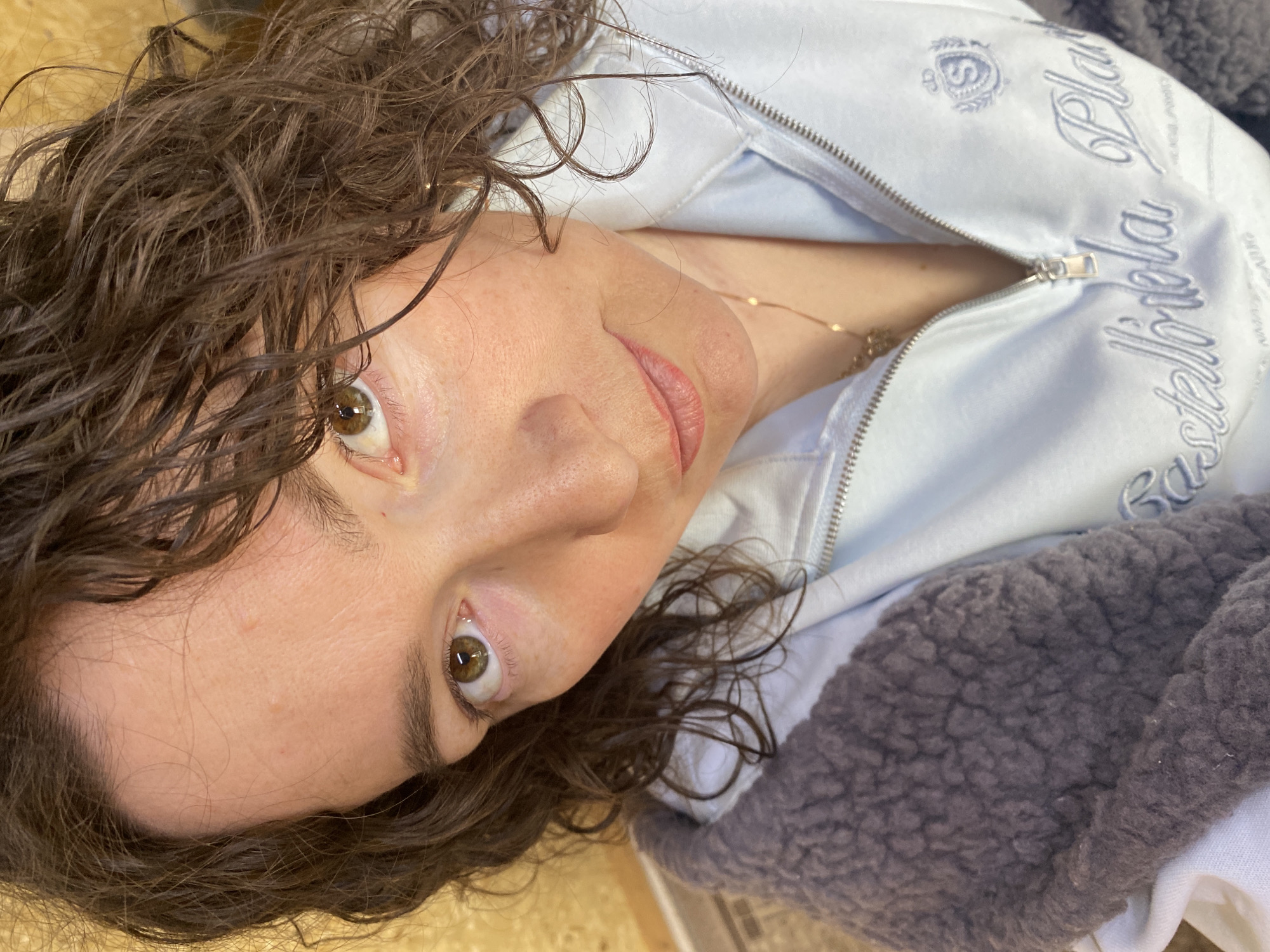A woman with brown curly hair and hazel eyes looks directly at the camera. She is wearing a light blue zip-up jacket with "Castelblola Pla" embroidered on it. A gold necklace is visible at the base of her neck. The background appears to be an interior setting. No indication of Ol'khovskoye, Russia is apparent.
