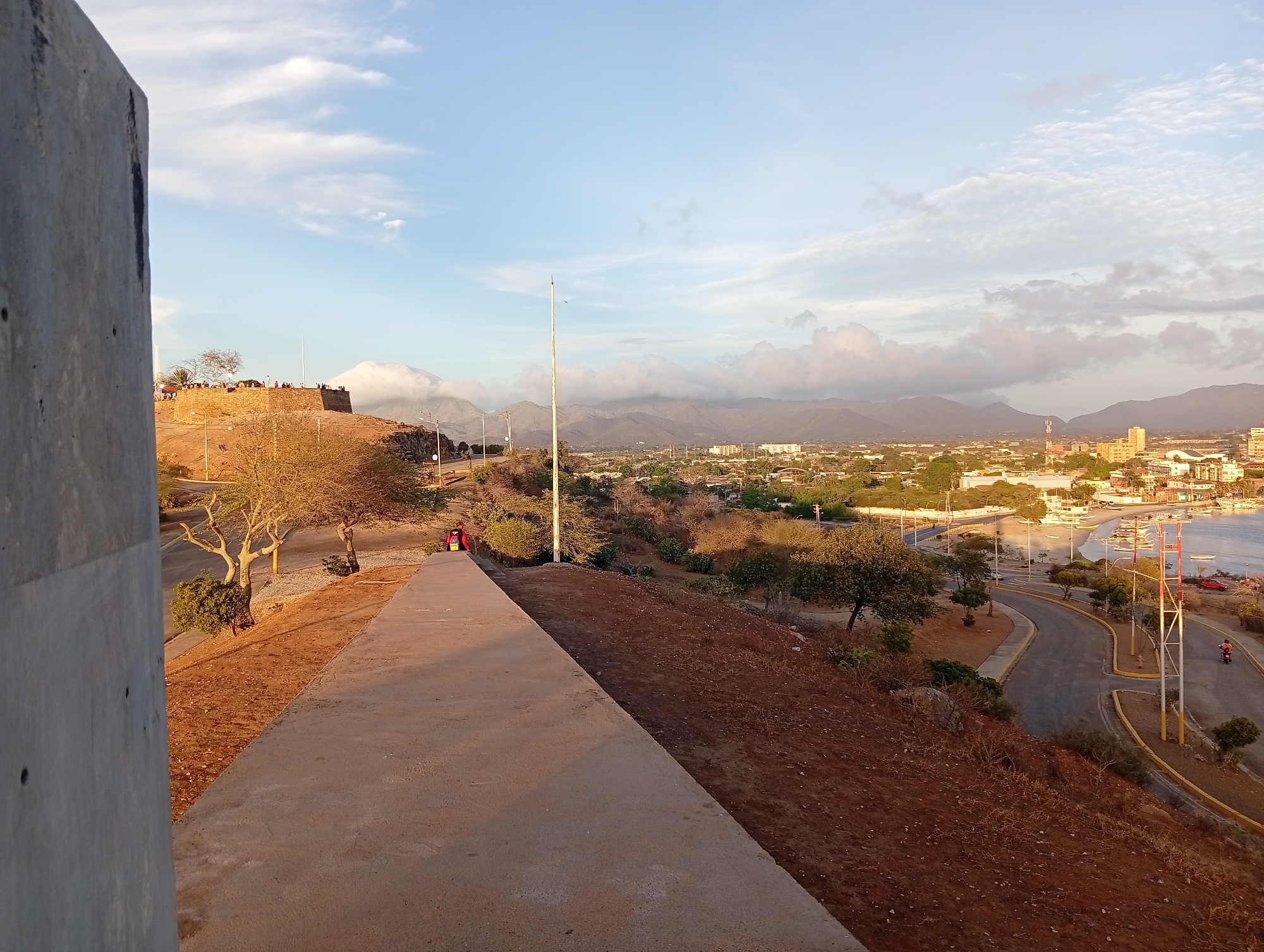 View of Juan Griego, Venezuela from a high vantage point. In the foreground is a concrete structure and a walkway. Beyond that, the hillside descends to a road that parallels a body of water with boats. The city of Juan Griego is visible in the middle ground, with buildings and streets. Mountains are in the background, partially obscured by clouds. A person is seen walking on the walkway.