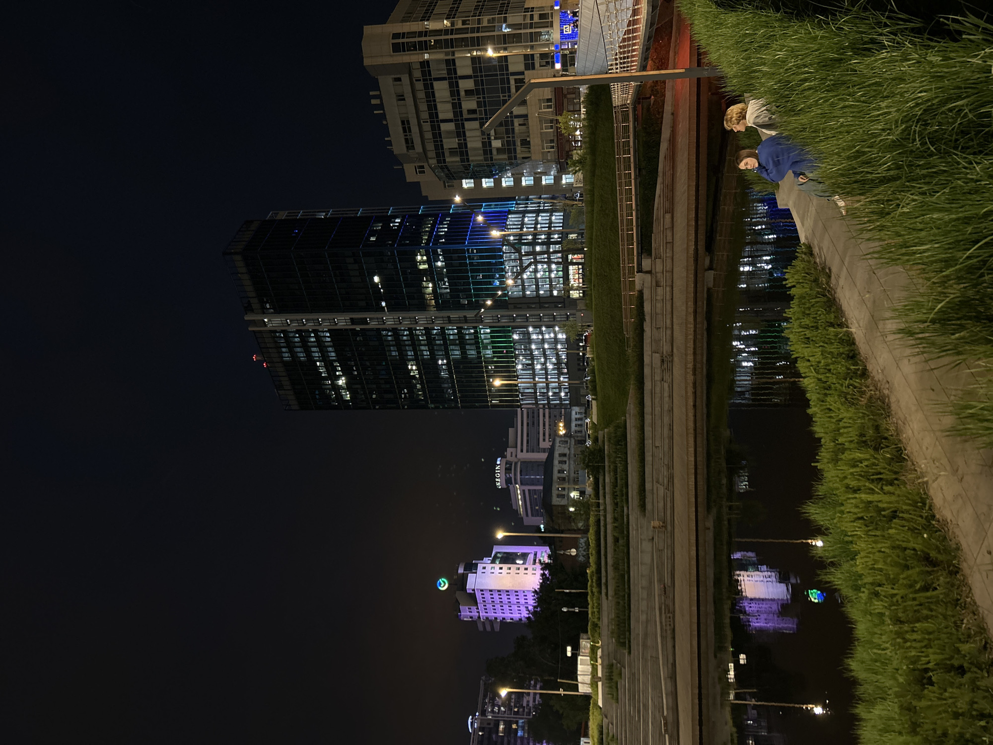 The image depicts a nighttime scene in Yekaterinburg, Russia, focusing on a waterfront area with modern architecture. A tall, dark skyscraper with illuminated windows dominates the right side of the frame, reflecting on the calm water in the foreground. To the left, a multi-story building is illuminated with vibrant purple and green lights, and a prominent circular green logo is visible at its peak. Adjacent buildings, some with patterned facades and lit windows, are also present.
A tiered, stepped embankment lines the water, with lush green grass growing along its edges. Two individuals, one wearing a blue hoodie and the other a light-colored top, are seated on the embankment, facing the water. They appear to be engaged in conversation or simply enjoying the view. Streetlights cast warm glows on pathways and bridges, contributing to the illuminated urban landscape. The sky is dark, indicating nighttime. The overall impression is one of a modern city park or public space at night, with a blend of natural elements and illuminated structures.