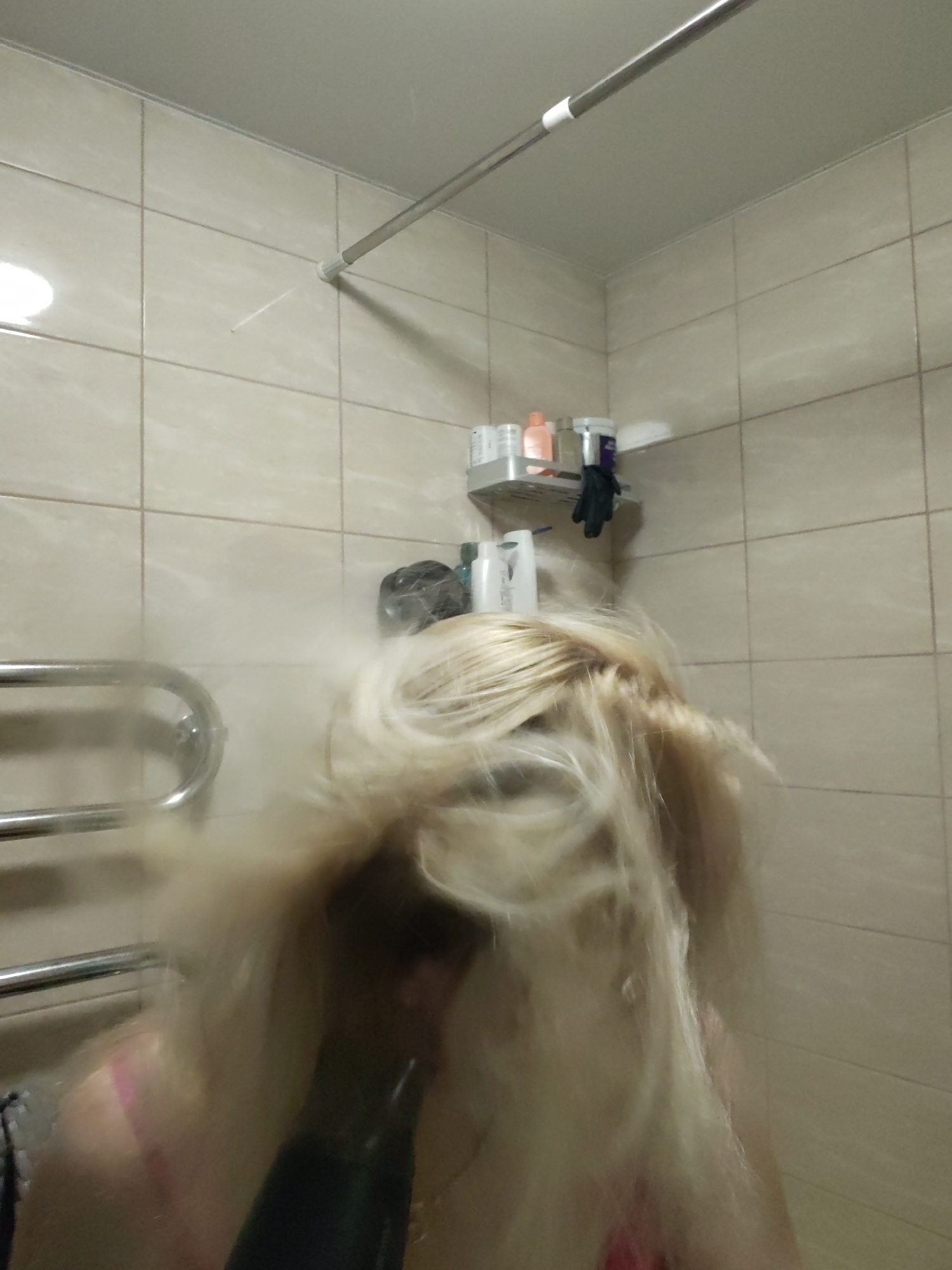 A blurry image shows a person with long, blonde hair using a black hairdryer in a bathroom. The bathroom is tiled with light beige square tiles and features a chrome towel rack on the left, a silver shower rod at the top, and a silver shelf with various bottles and a pair of black gloves on the wall. The exact location within Izhevsk, Russia is impossible to determine from the image.