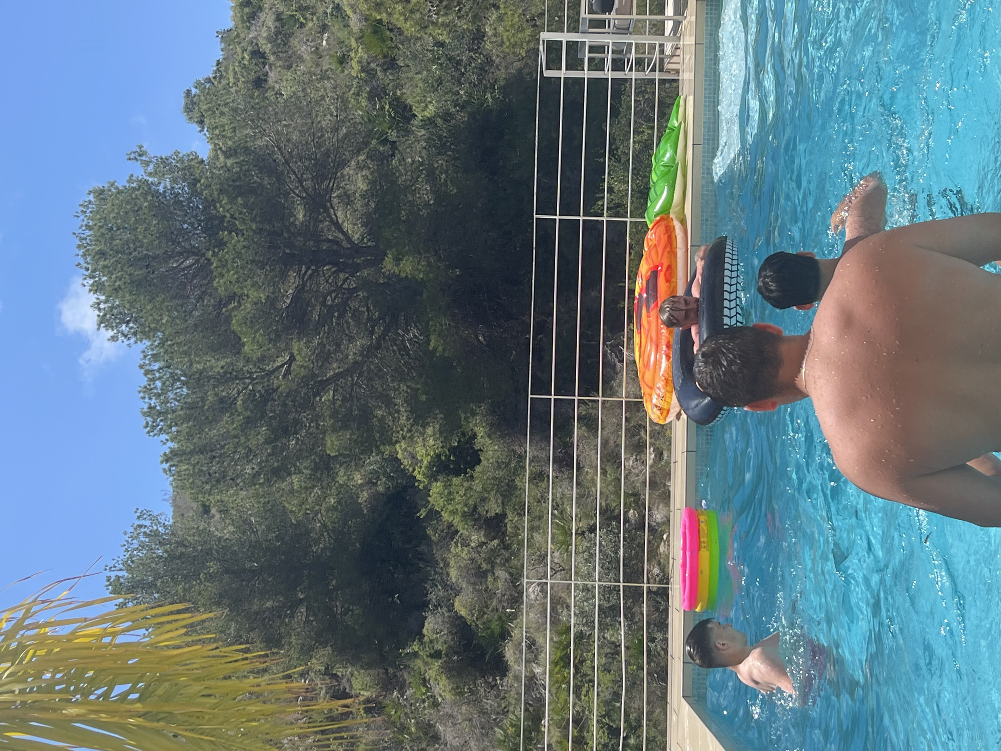 The image shows a group of people in a swimming pool in El Poble Nou de Benitatxell / Benitachell, Spain. The pool is surrounded by a metal railing, beyond which is a densely wooded area and a hillside. Some inflatable toys, including a pineapple, a green alligator, and a colorful ring, are near the edge of the pool. Two adults are near the edge of the pool. One boy is swimming in the pool. The sky is blue.