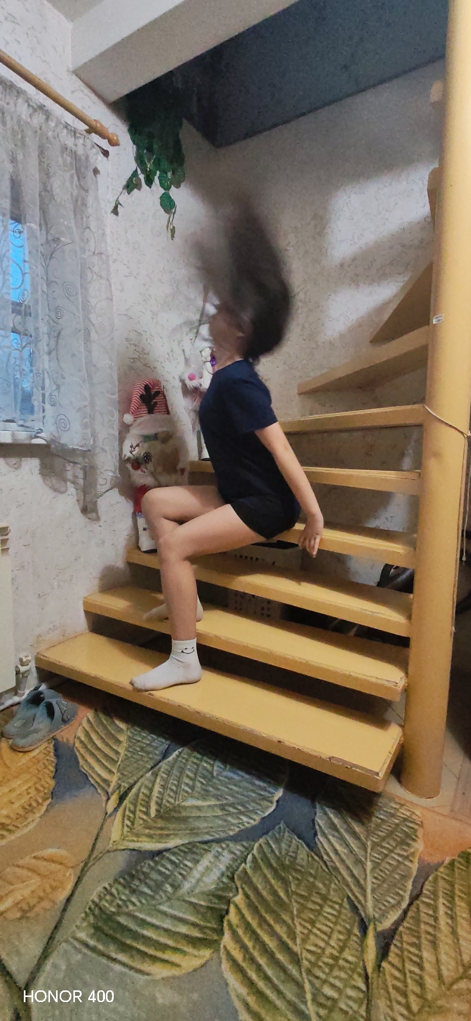 The photograph shows a person with dark hair that is blurred, likely due to motion. The person is sitting on wooden stairs with a yellow tint. They are wearing a dark blue t-shirt and dark shorts. On the person's feet are white socks, one of which has a smiley face on it. Behind the person, to the left, is a window with patterned curtains. There are also some stuffed animals visible. To the right, the stairs spiral upwards, supported by a yellow post. The floor covering below the stairs has a large leaf pattern. In the bottom left corner, there's a faint logo for "HONOR 400." The location is identified as Noviy, Russia.