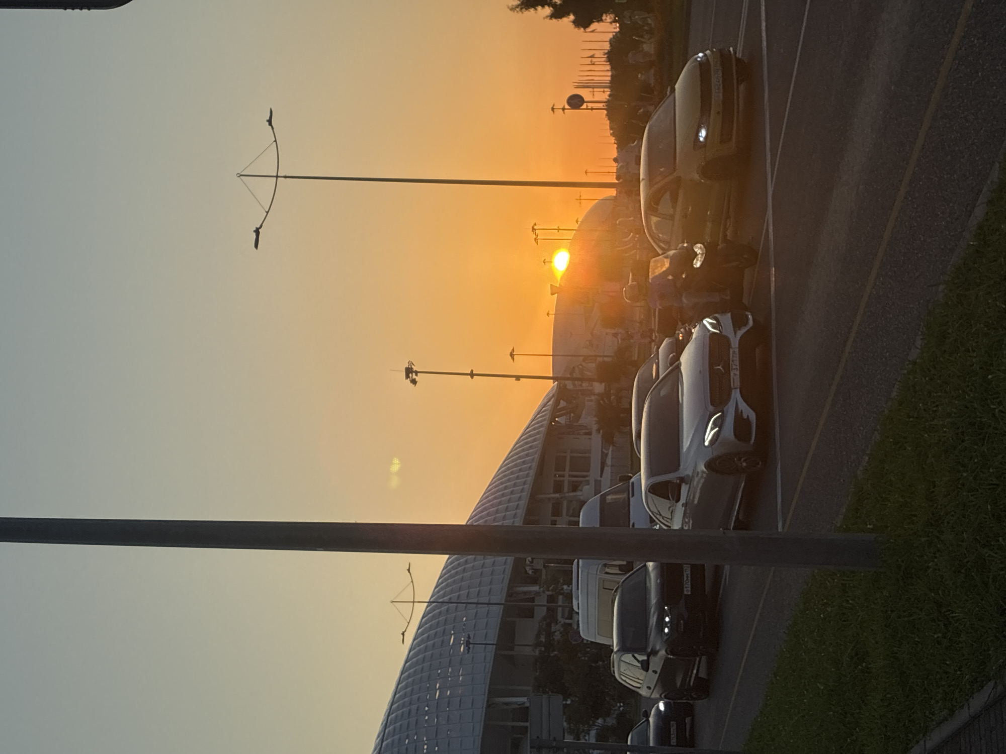 The image captures a sunset scene in the city of Sirius, Russia, near the Olympic Park. The sky displays a gradient of warm colors, transitioning from a soft yellow near the horizon to a pale blue overhead. The sun, a bright orb, is setting behind a large, modern, white-domed structure, possibly a stadium or exhibition center, casting a golden glow over the scene.
In the foreground, a multi-lane road is visible with several vehicles. A dark-colored SUV is in the left lane, followed by a white van. A light-colored sedan is in the adjacent lane, with a motorcycle carrying a rider in a blue and white outfit positioned to its right. Further to the right, a yellow sedan is also on the road. The vehicles appear to be in traffic, suggesting a common occurrence during peak hours or due to road construction.
Streetlights with multiple lamps stand tall along the roadside, some silhouetted against the vibrant sky. The area around the road is lined with grass, and some trees are visible in the distance, adding to the urban landscape. The architecture of the dome-like structure is modern, characterized by its curved roof and large windows. The overall atmosphere is one of a typical evening commute in a modern, well-developed urban area, bathed in the dramatic light of a setting sun.