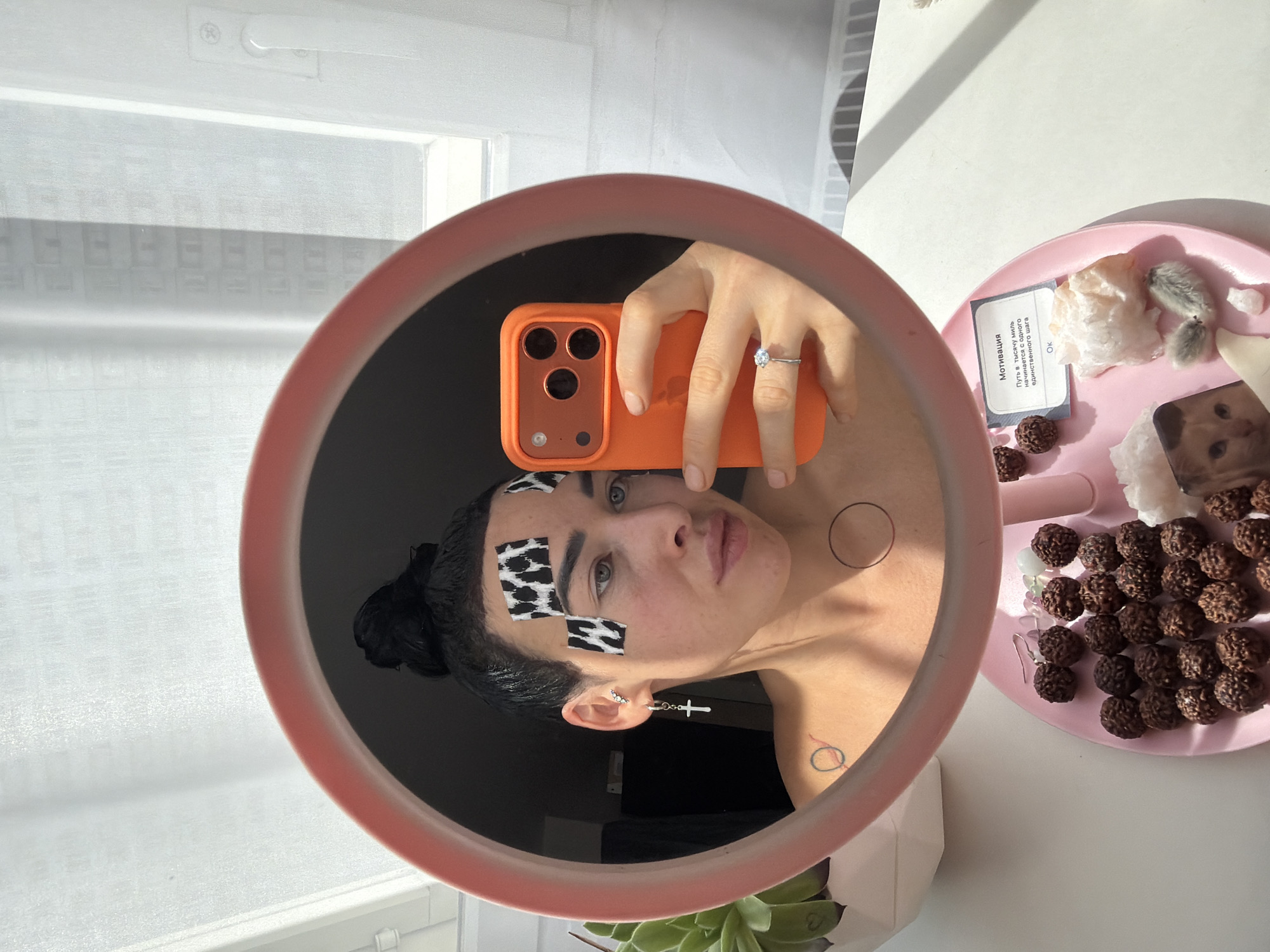 A person is taking a mirror selfie in an indoor setting, likely during the daytime given the bright natural light. The central subject is the reflection of the person in a round mirror with a pinkish-orange frame. The person has dark hair pulled back and is looking directly into the camera of an orange-cased smartphone held in their hand, which is also visible in the mirror. They are wearing a ring on their left ring finger and a cross earring on their right ear. Several cow-print patches are applied to their forehead and cheek. Two small, simple circular tattoos or drawn symbols are visible on their collarbone/shoulder area.
To the right of the mirror, a light pink tray holds a collection of items, including many small, dark, brown, beaded objects (possibly Rudraksha beads), several light-colored, irregular crystals or stones, two fuzzy decorative objects, and a small square block or coaster featuring a cat's face. A small card with Russian text is also on the tray. The text reads: "МОТИВАЦИЯ. Путь в тысячу миль начинается с одного единственного шага. ОК." (MOTIVATION. A journey of a thousand miles begins with a single step. OK.)
Part of a green succulent plant in a white pot is visible at the bottom edge of the image, outside the mirror's frame. The background reflected in the mirror shows a bright space with white walls and a window covered by sheer white curtains.