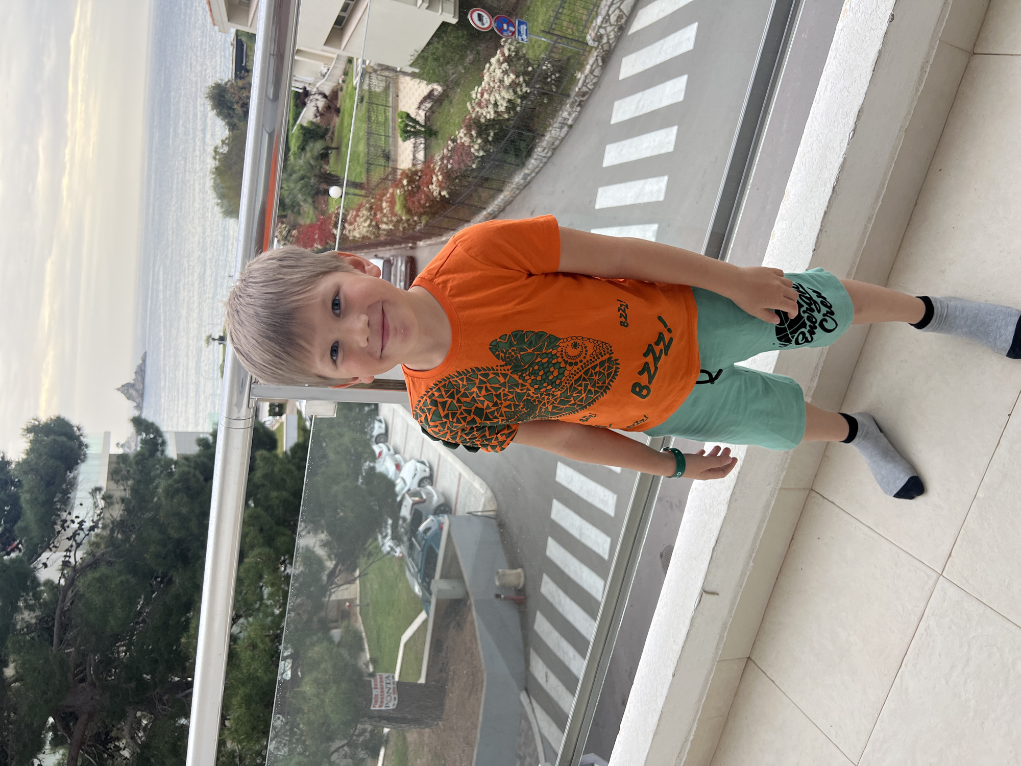 A young boy stands on a balcony overlooking the Adriatic Sea in Petrovac centar, Montenegro. He wears an orange t-shirt with a lizard design, green shorts, and grey socks with black toes. The balcony has a glass railing. A street with a zebra crossing and parked cars is visible below. Trees and buildings frame the scene. The sky is overcast.
