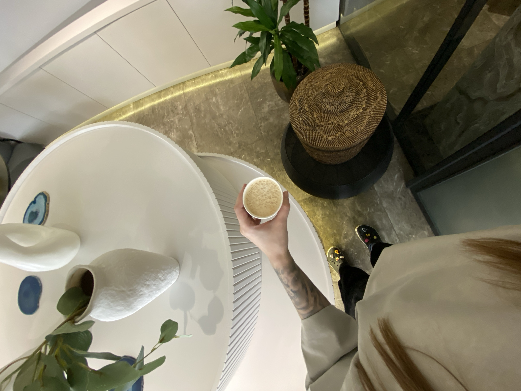 A top-down shot depicts a person holding a cup of coffee in a room with modern decor. The person, visible from the torso down, wears black pants, sandals adorned with stickers, and a light-colored shirt or sweater. They hold a white cup filled with a beige, foamy liquid, presumably coffee or a similar beverage.
The coffee cup is held above a white, circular table with a ribbed edge. On the table, there is a white vase with green foliage inside and decorative elements such as blue circles and amorphous white shapes. Nearby is a woven basket sitting atop a dark circular stand. A potted green plant is situated near the basket.
The floor is light brown with what appears to be tile or stone pattern. There is a golden light strip along the wall, and a dark-framed glass door or window is visible to the right.
There is no indication in the image as to whether this scene is in Votkinsk, Russia.